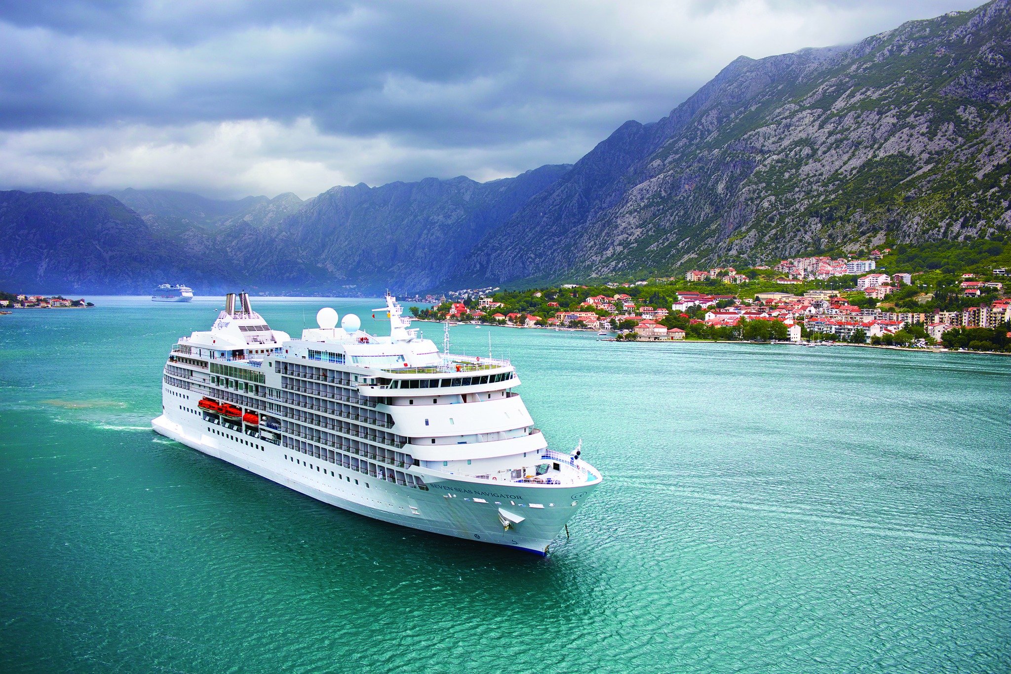 Large white cruise ship sailing on a body of water with greenish-blue color, with a town featuring red-roofed buildings along the shoreline and steep mountainous terrain in the background under cloudy skies.
