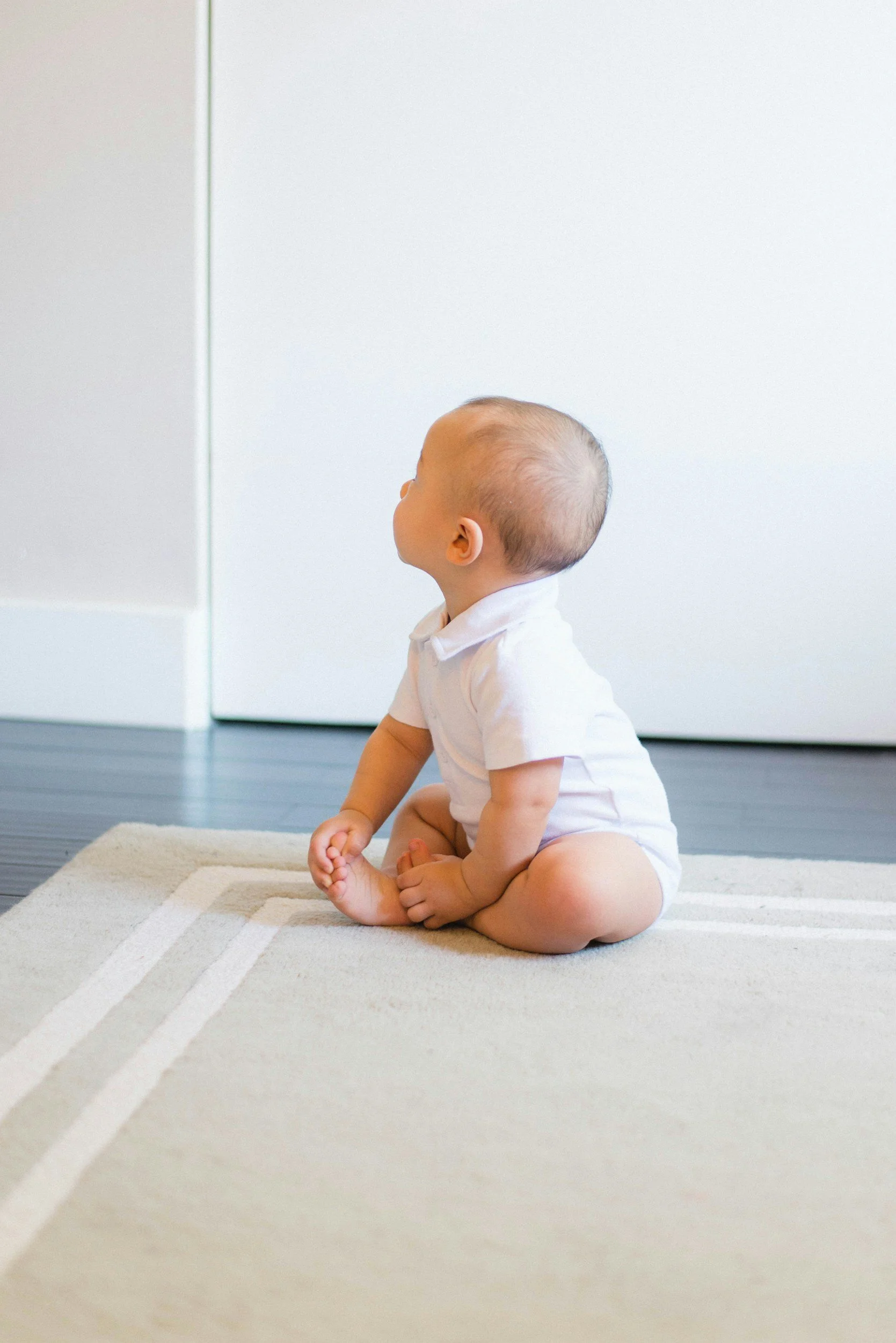 A young baby with a shaved head sitting on a patterned beige rug, looking to the left, surrounded by white walls and a white cabinet.