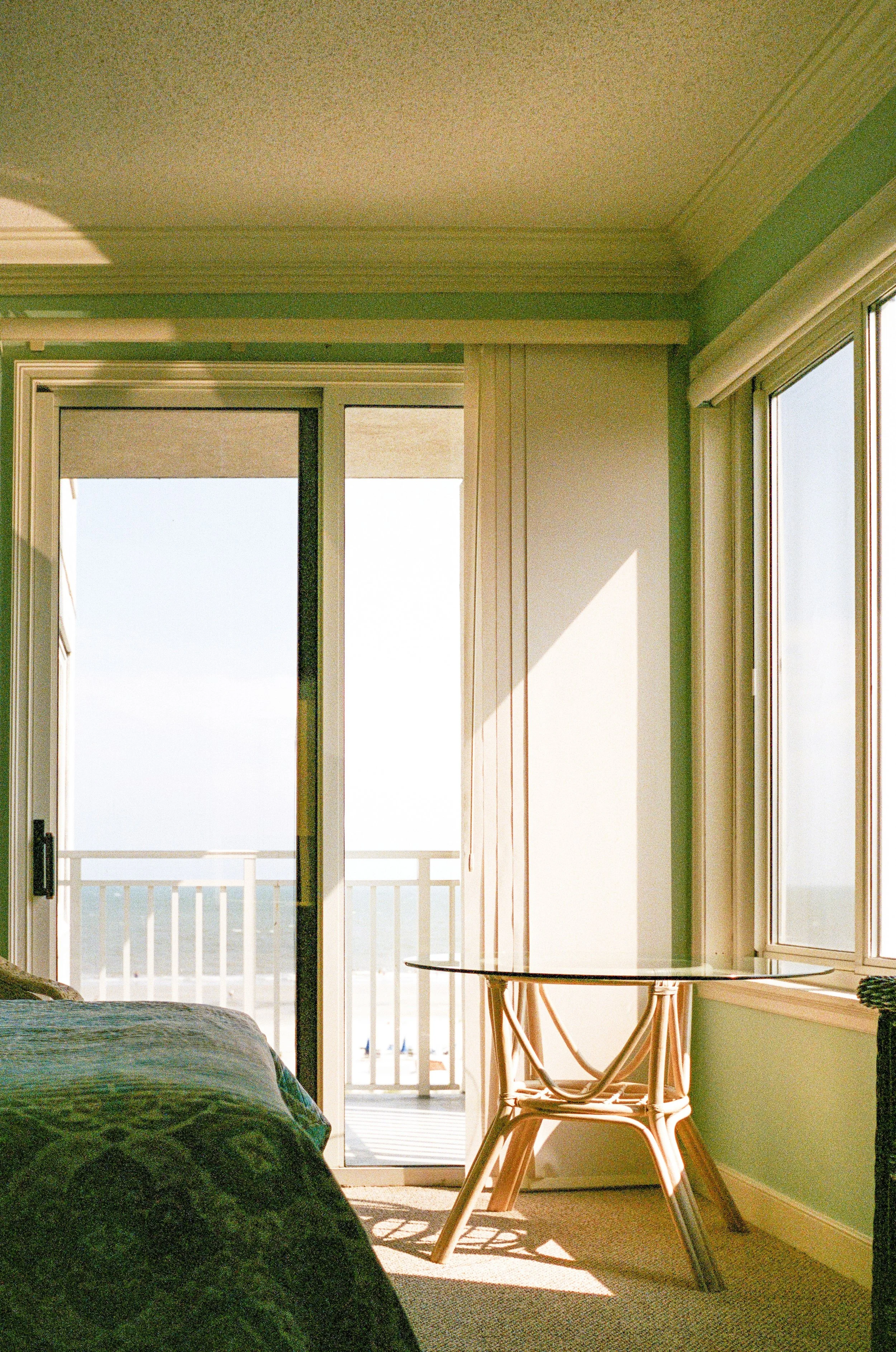 Sunlit hotel room with sliding glass door leading to balcony, wooden chair near window, and bed partially visible in lower corner.