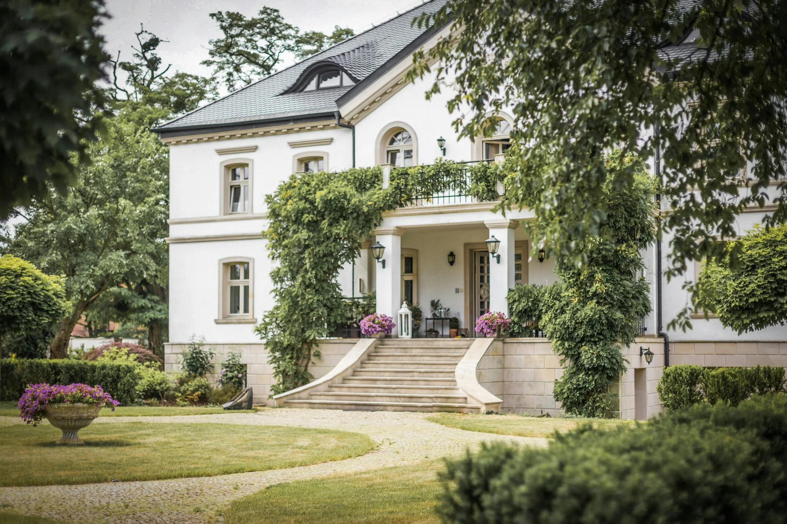 A large white house with a dark roof, two curved staircases leading to the front entrance, and green vines growing on the exterior walls. The house is surrounded by a landscaped lawn with trees, bushes, and potted flowers.