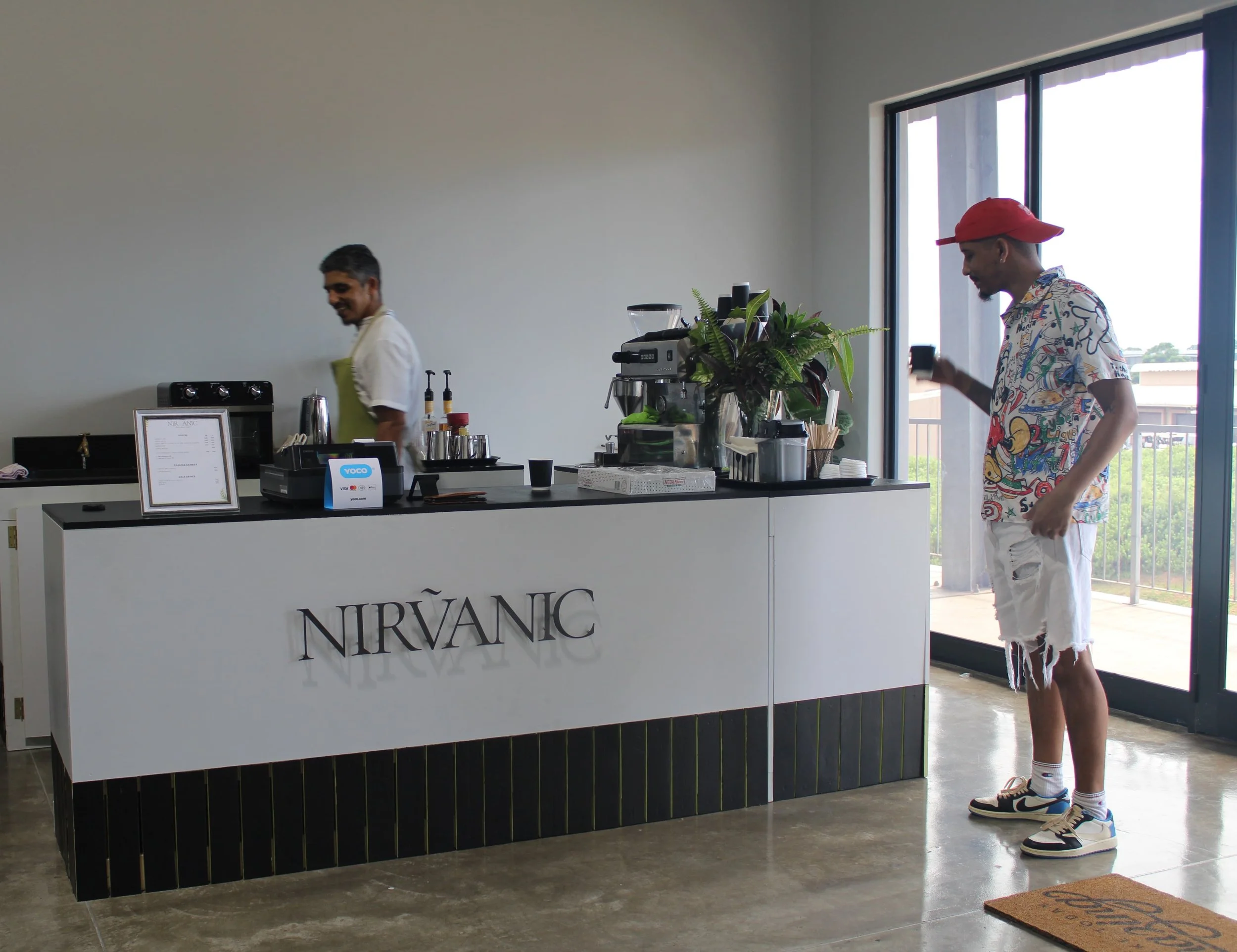 A customer in casual clothing and a red cap is standing at a coffee bar, using a smartphone. The barista behind the counter is smiling and working, with coffee equipment and a plant on the counter. The coffee shop has a sign reading 'NIRVÂNIC' and la