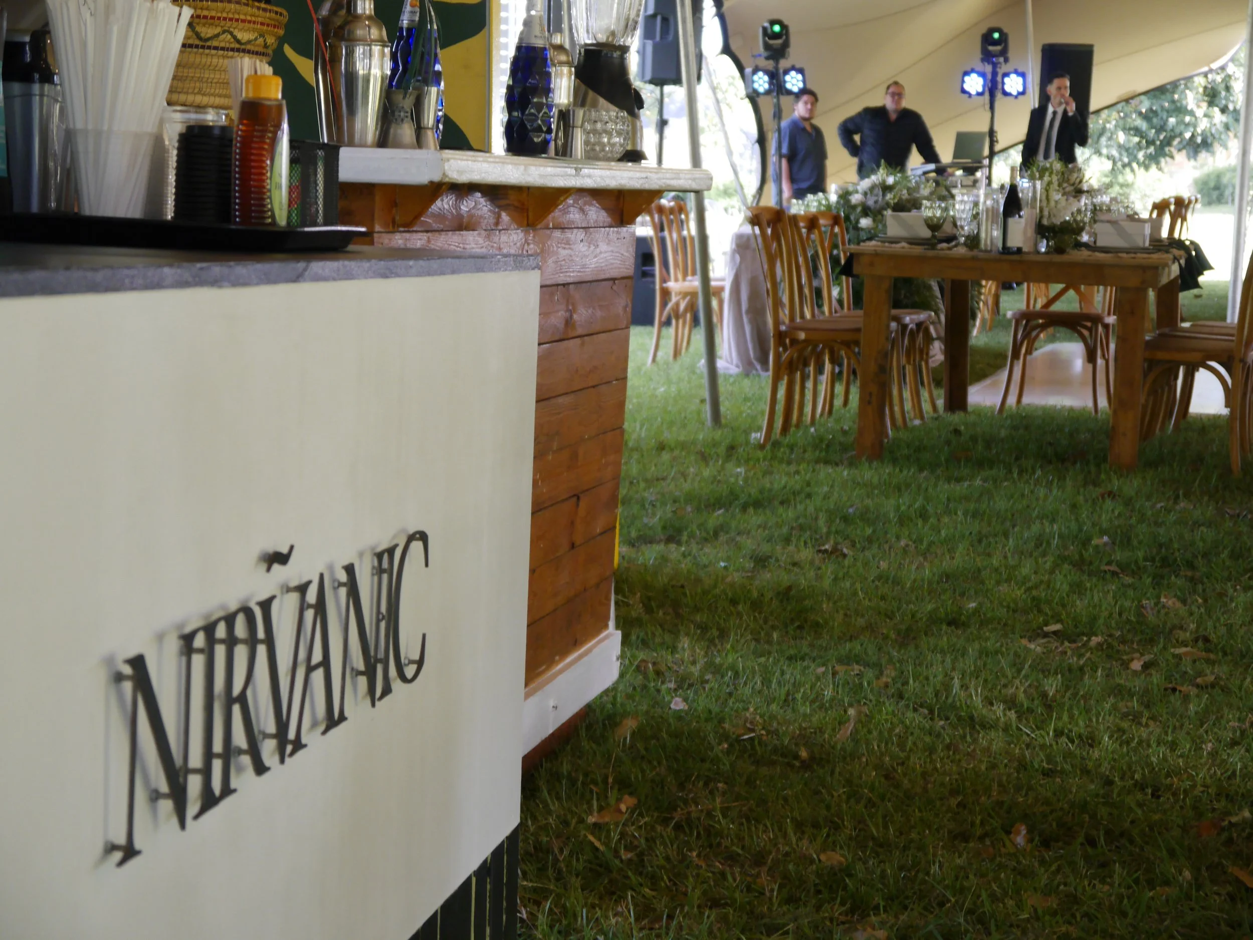 Wedding reception setup with round tables and wooden chairs under a tent, with a bar area in the foreground featuring condiments and drinkware, and a DJ booth with lights and people in the background.