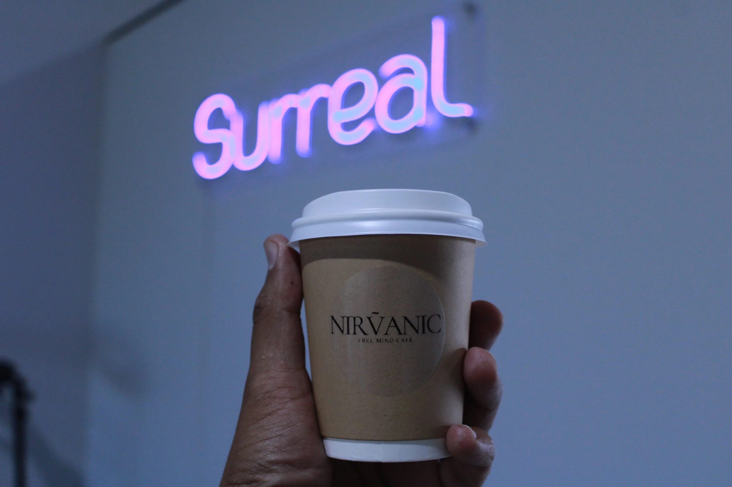 Hand holding a disposable coffee cup with a beige sleeve that has 'NIRVĀNIC FREE MIND CAFE' printed on it. In the background, there is a purple neon sign that reads 'surreal' on a gray wall.
