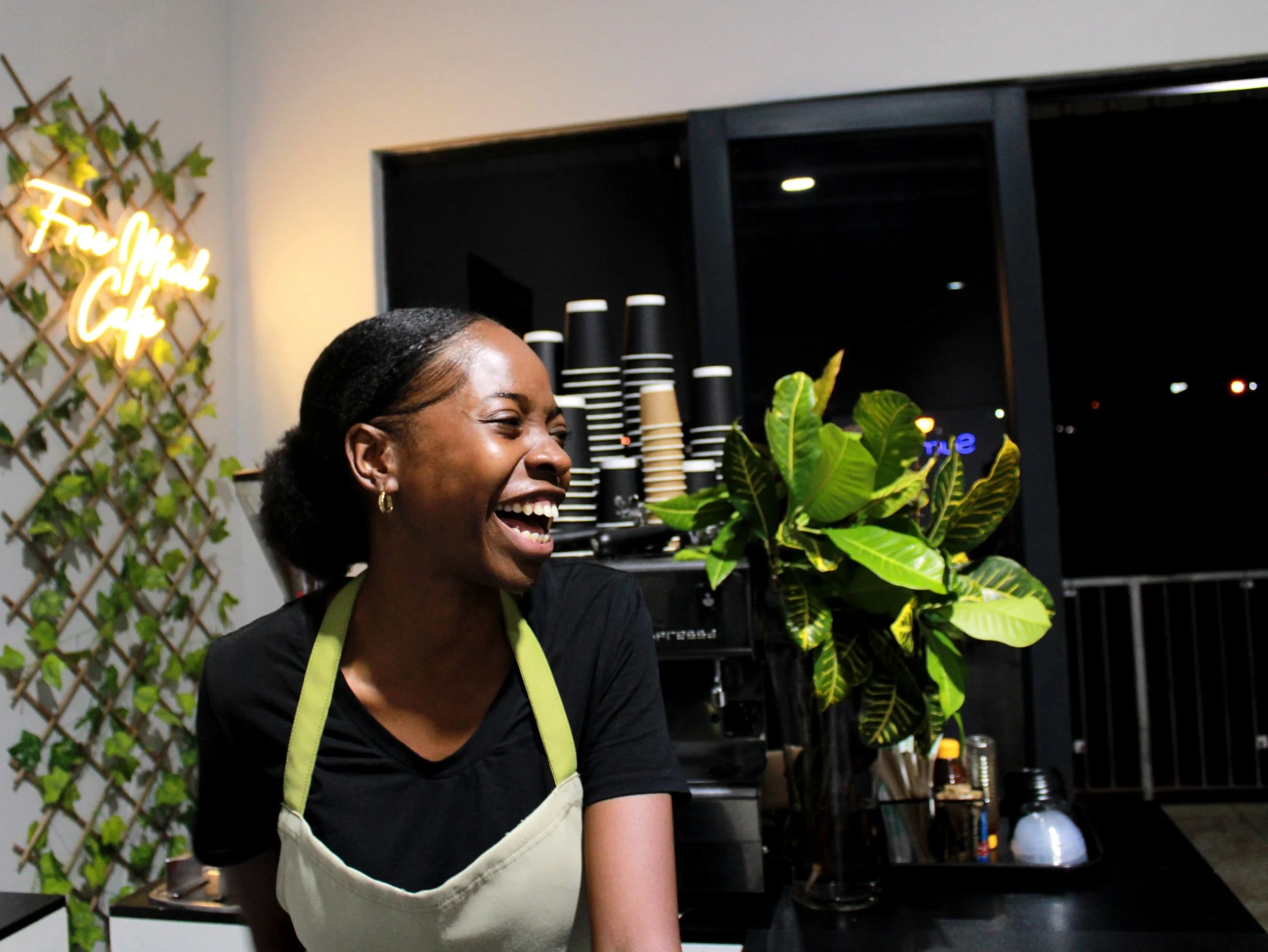 A smiling woman in a black shirt and apron, standing inside a cafe, with a sign that reads 'Free Mind Cafe' and a large green plant on a counter behind her.