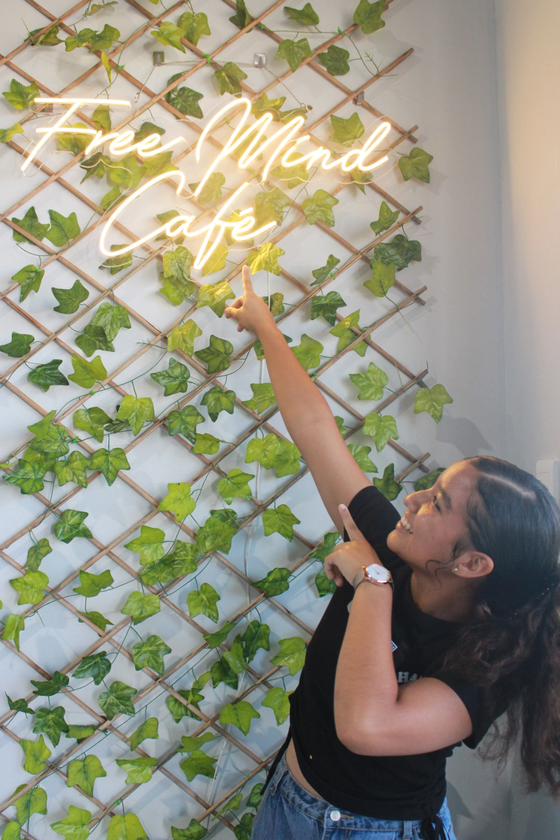 Woman smiling and pointing at a neon sign that says 'Free Mind Cafe', with a decorative wall of faux green ivy and a wooden lattice in the background.