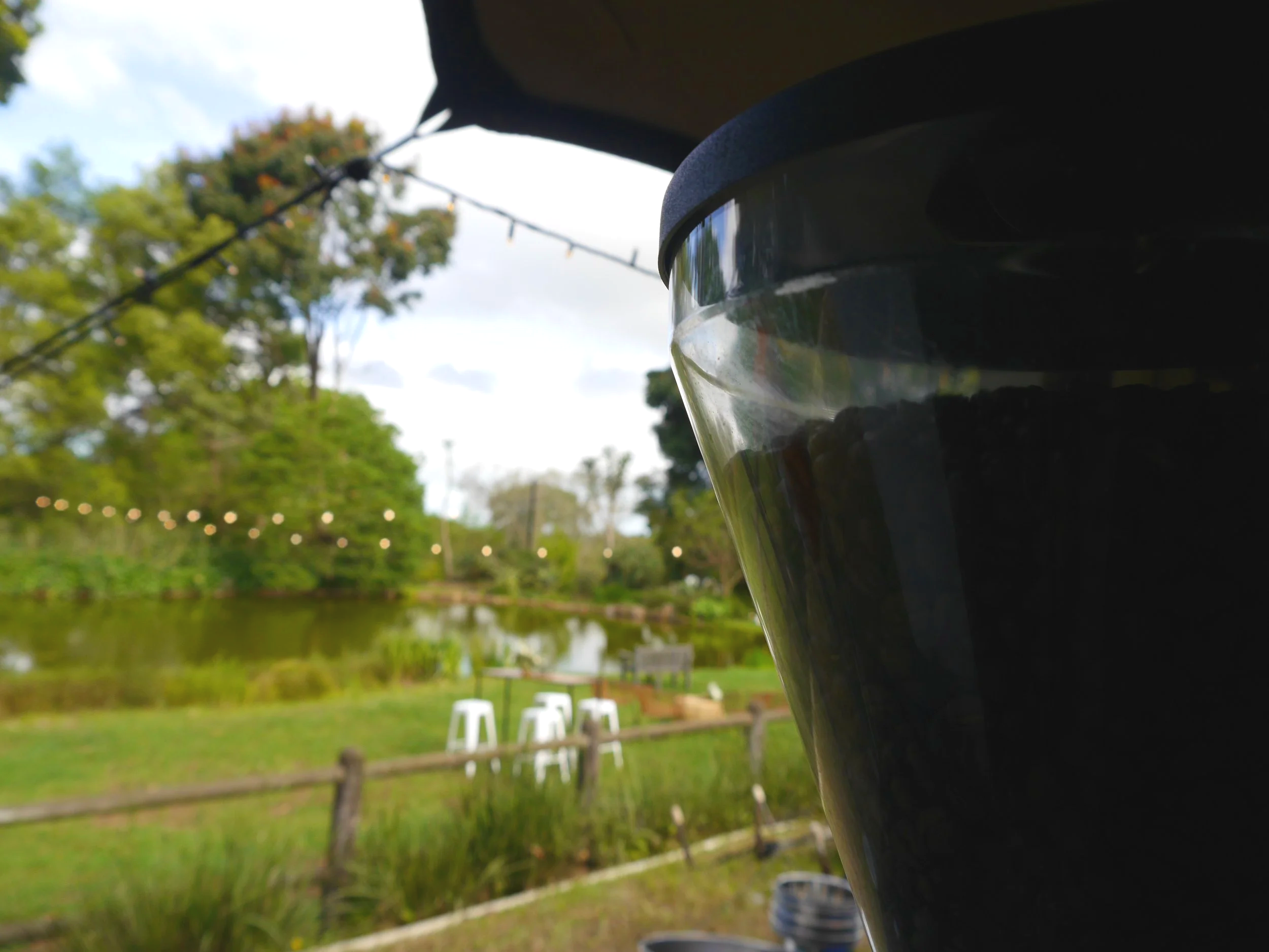 Close-up of a black coffee cup outdoors with a pond, trees, string lights, and outdoor furniture in the background.