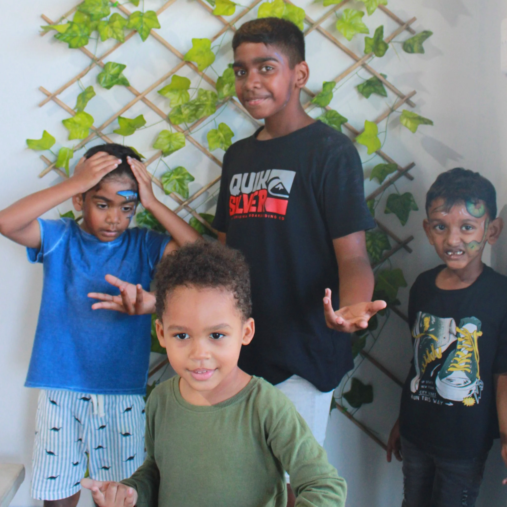 Four boys standing in front of a leafy background, showing different facial expressions and hand gestures.