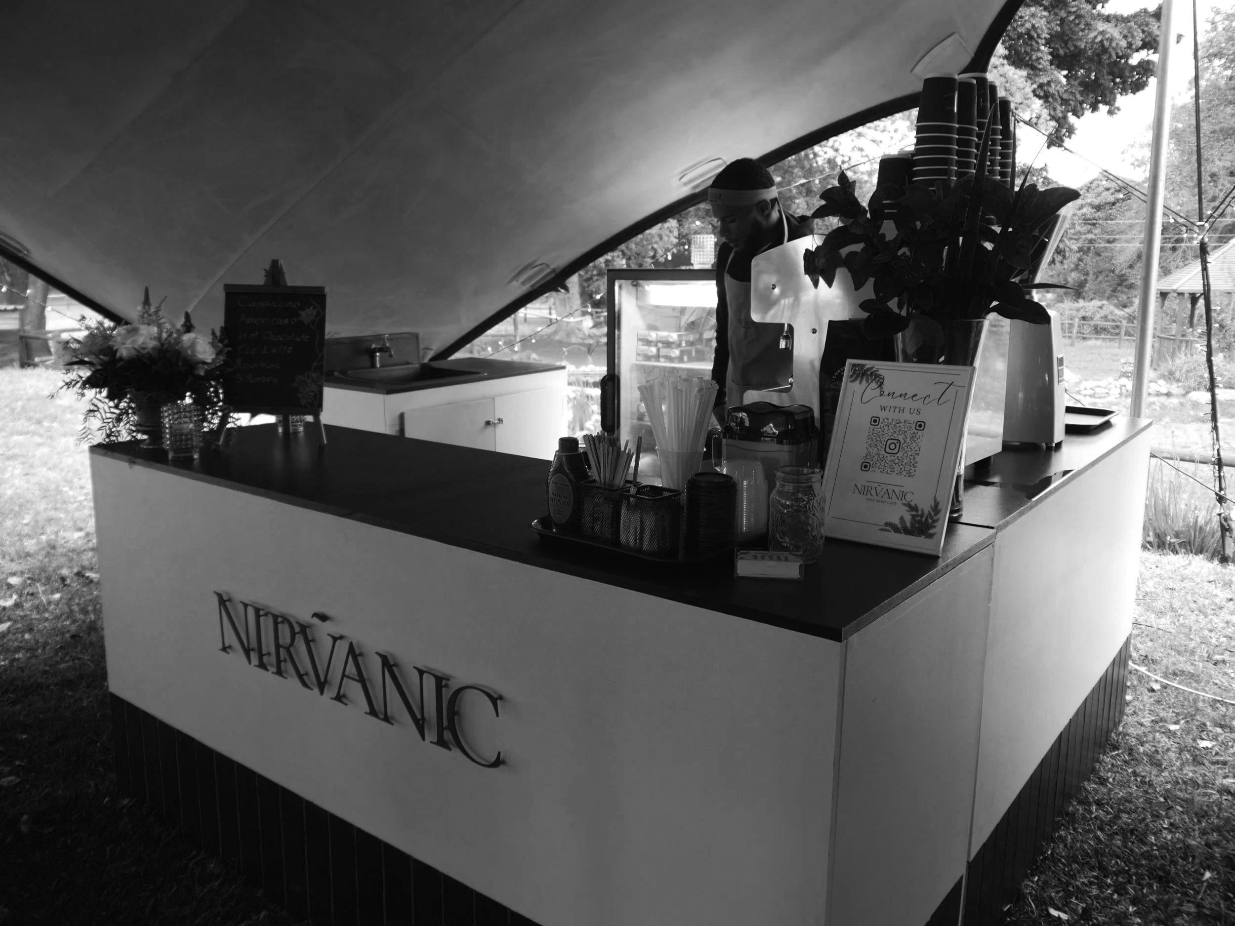 A beverage stand that has the name 'Nirvanic' on the front, set up outdoors under a tent, with a person preparing drinks behind the counter and various supplies on the counter.