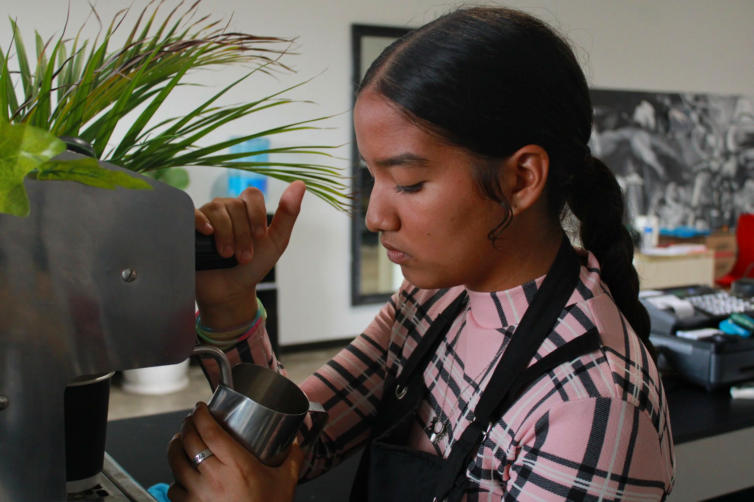 A young woman with dark hair tied back in a ponytail adds milk to coffee behind a coffee machine, with a potted plant nearby and a busy office environment in the background.