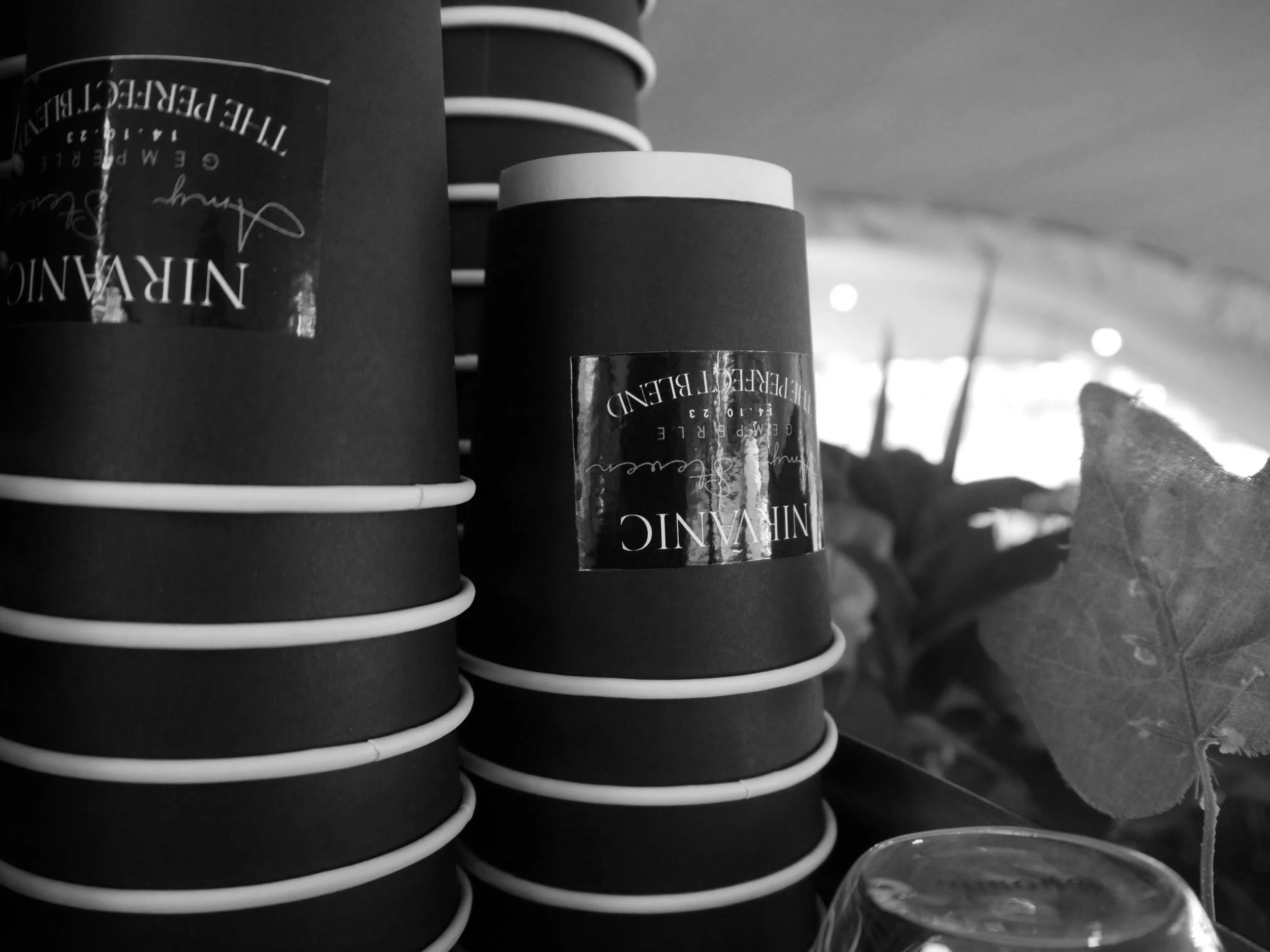 Stacked black cups with a label reading "NIRVANIC" and the words "THE PERFECT BLEND." There is a leaf and a glass in the foreground.