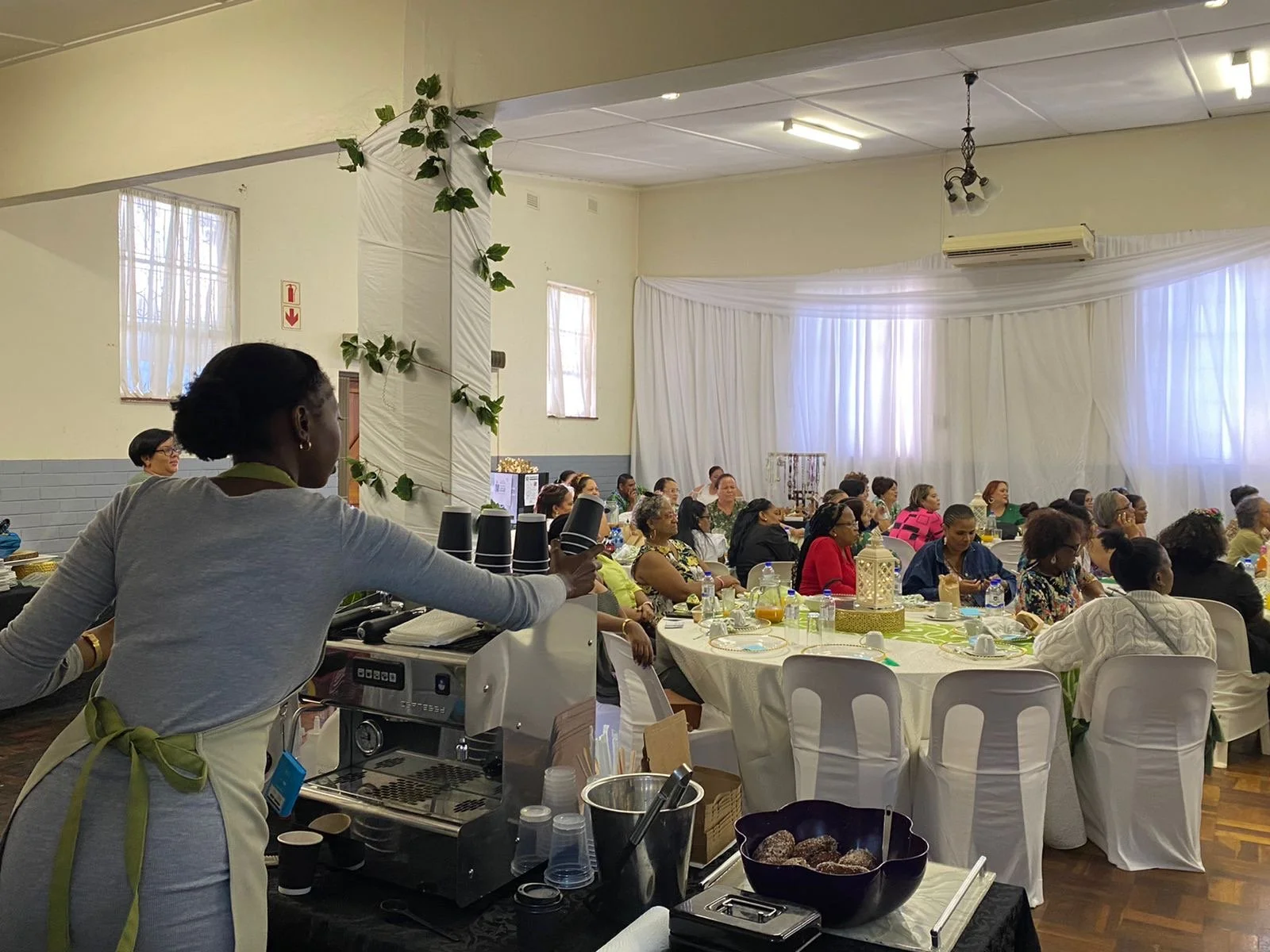 Women seated at tables during a meeting or seminar in a decorated hall with white drapery and green accents.