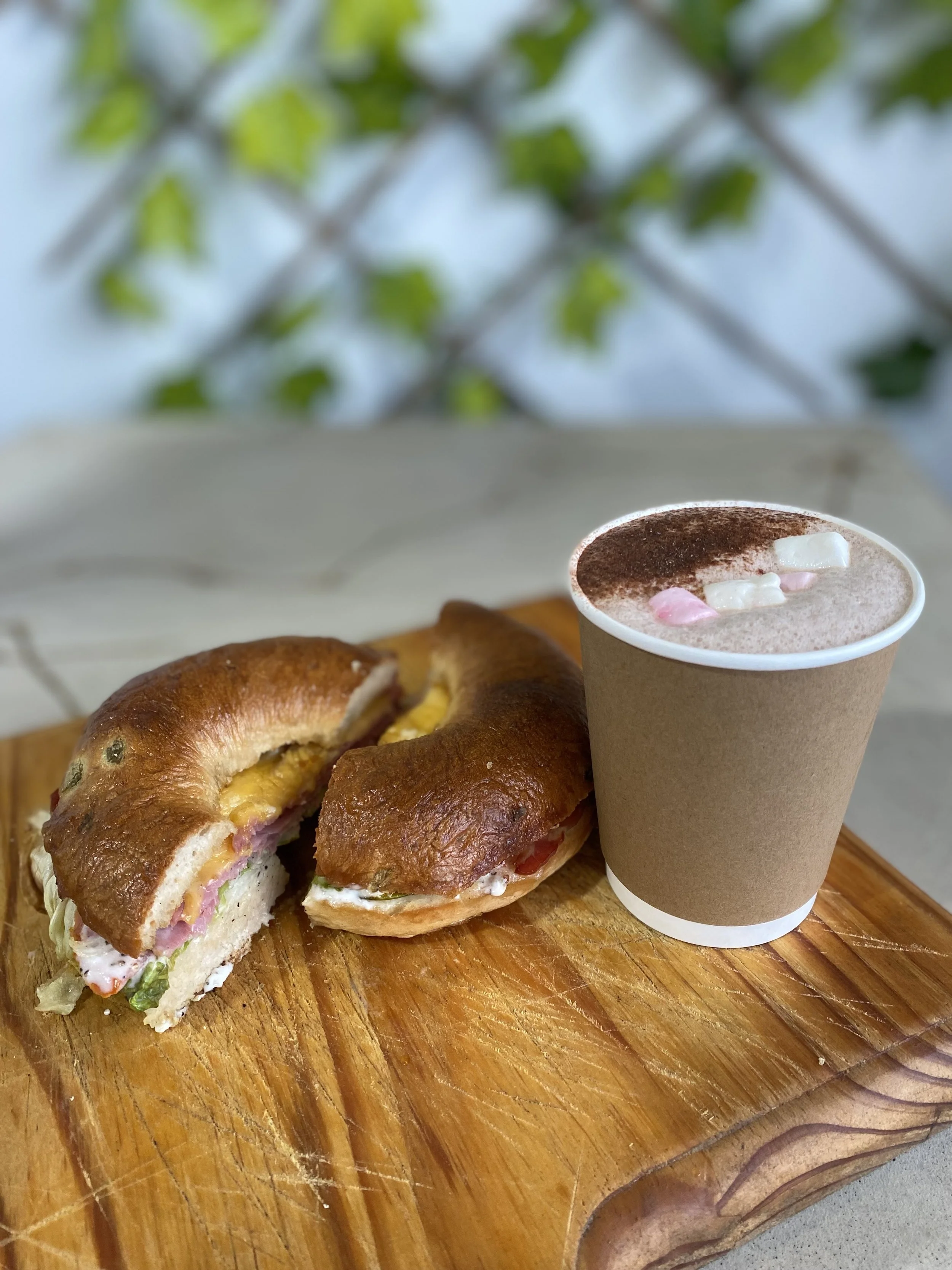 A croissant sandwich with ham, cheese, and vegetables on a wooden serving board, accompanied by a paper cup of hot cocoa topped with marshmallows.