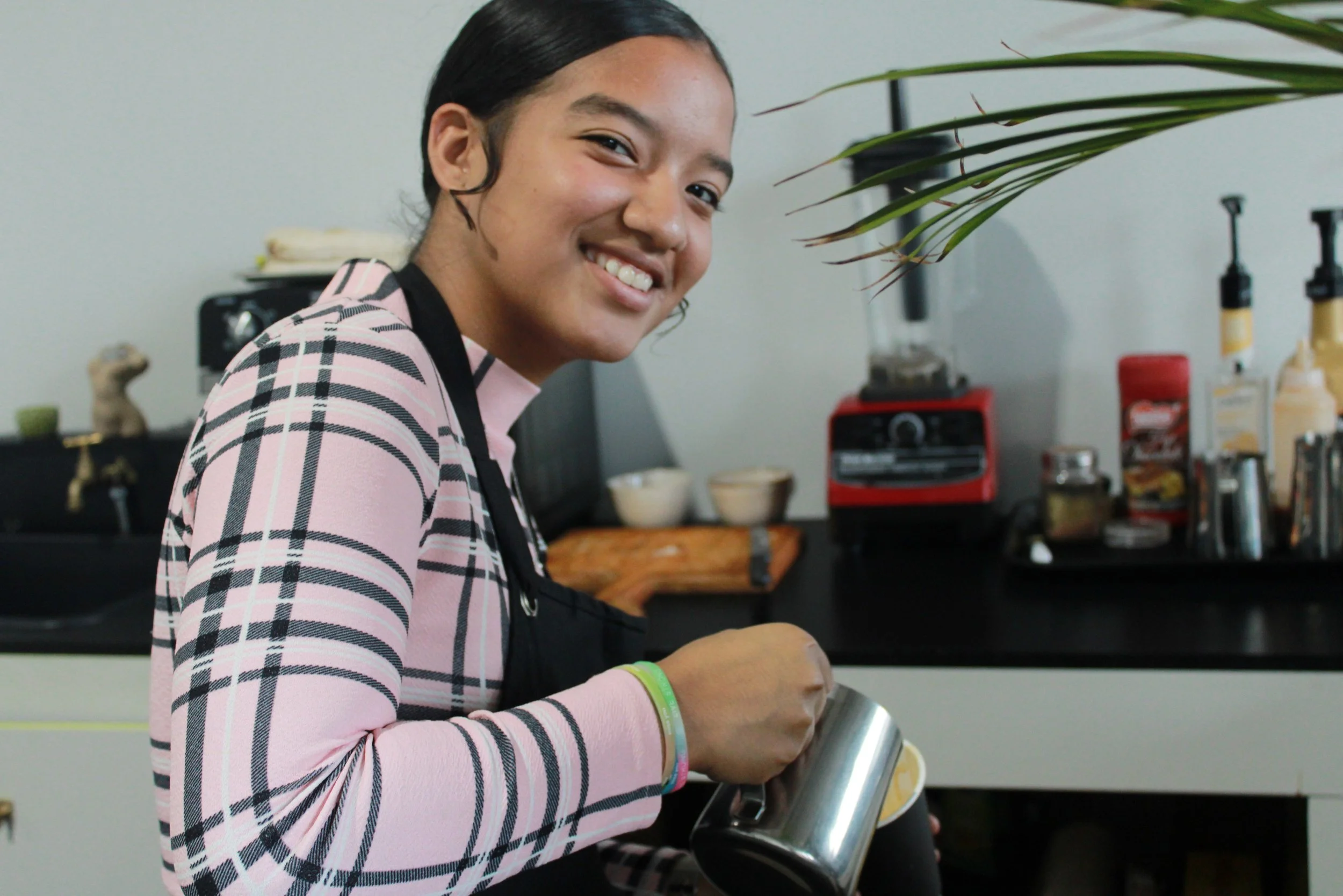 A young woman with black hair in a ponytail, smiling while holding stainless steel in a modern kitchen. She wears a pink and black plaid long-sleeve shirt and a black apron, with a green bracelet on her wrist. The kitchen has a black counter with var