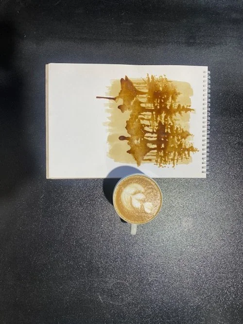 A sketchbook with a landscape drawing, and a cup of coffee with latte art on a black table.