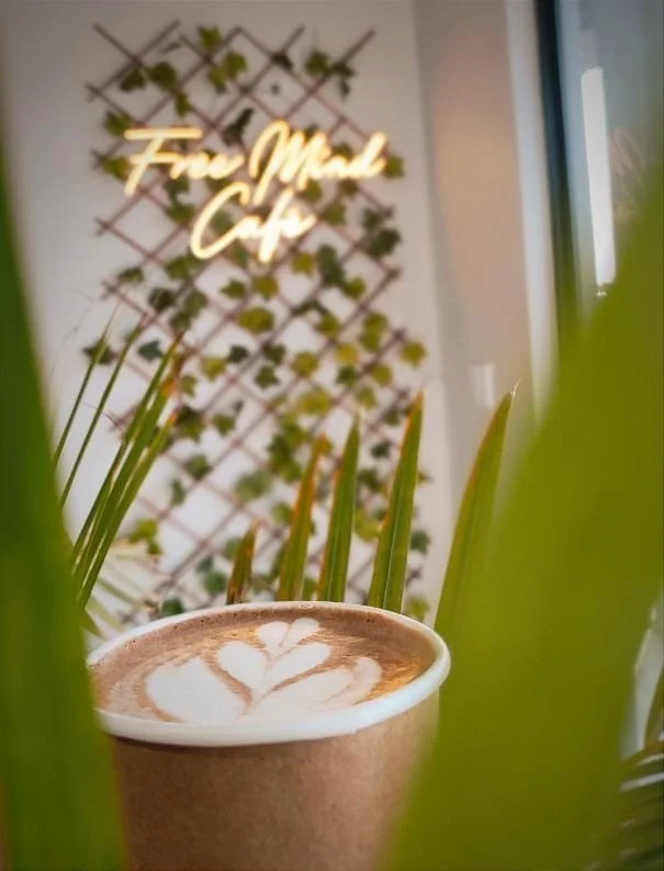 A cup of coffee with latte art on top, placed on a surface with plant leaves partially obscuring the view. In the background, there's a wall with a neon sign that reads 'Free Mind Cafe', and a wall decorated with a lattice with small green plants.
