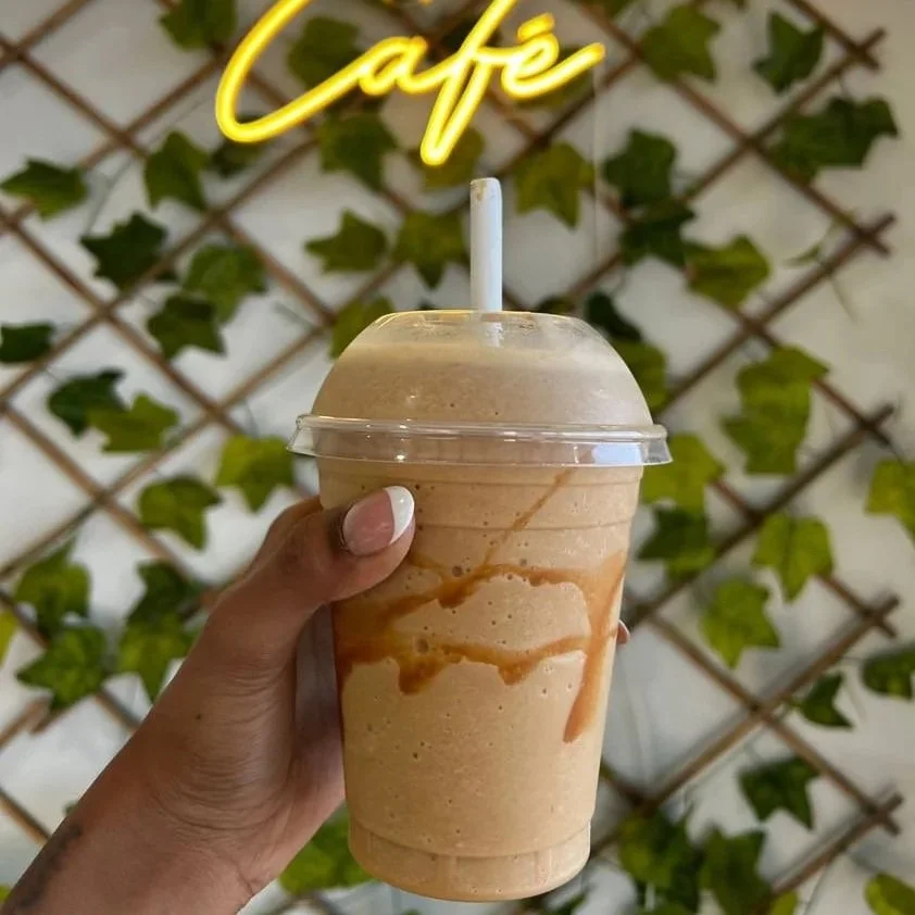 Person holding a blended coffee drink with caramel drizzle in a clear plastic cup with a lid and a straw, in front of a wall decorated with green ivy and a yellow neon 'Café' sign.
