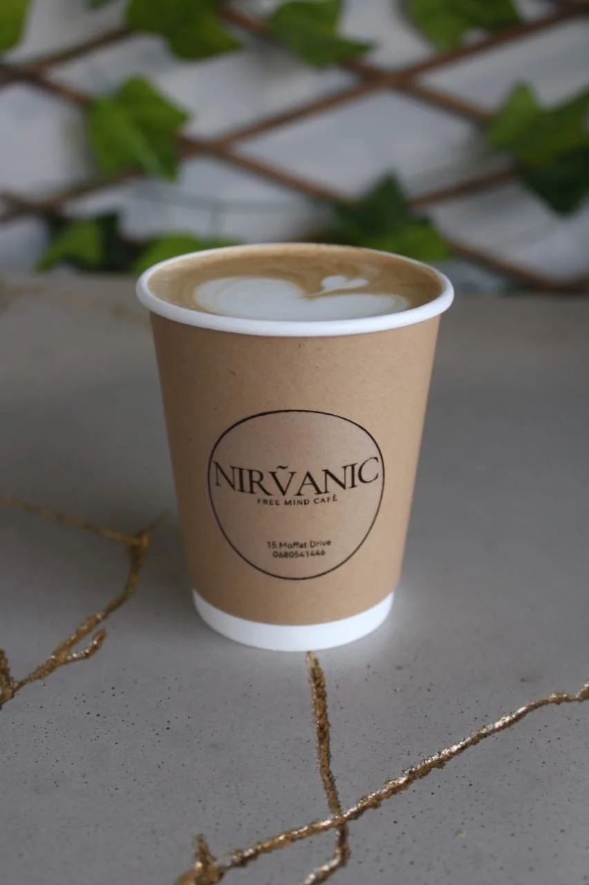 A paper coffee cup with the label 'NIRVĀNIC FREE MIND CAFÉ' sitting on a handmade concrete table some decorative gold cracks, and a blurred background with green leaves and a white wall.