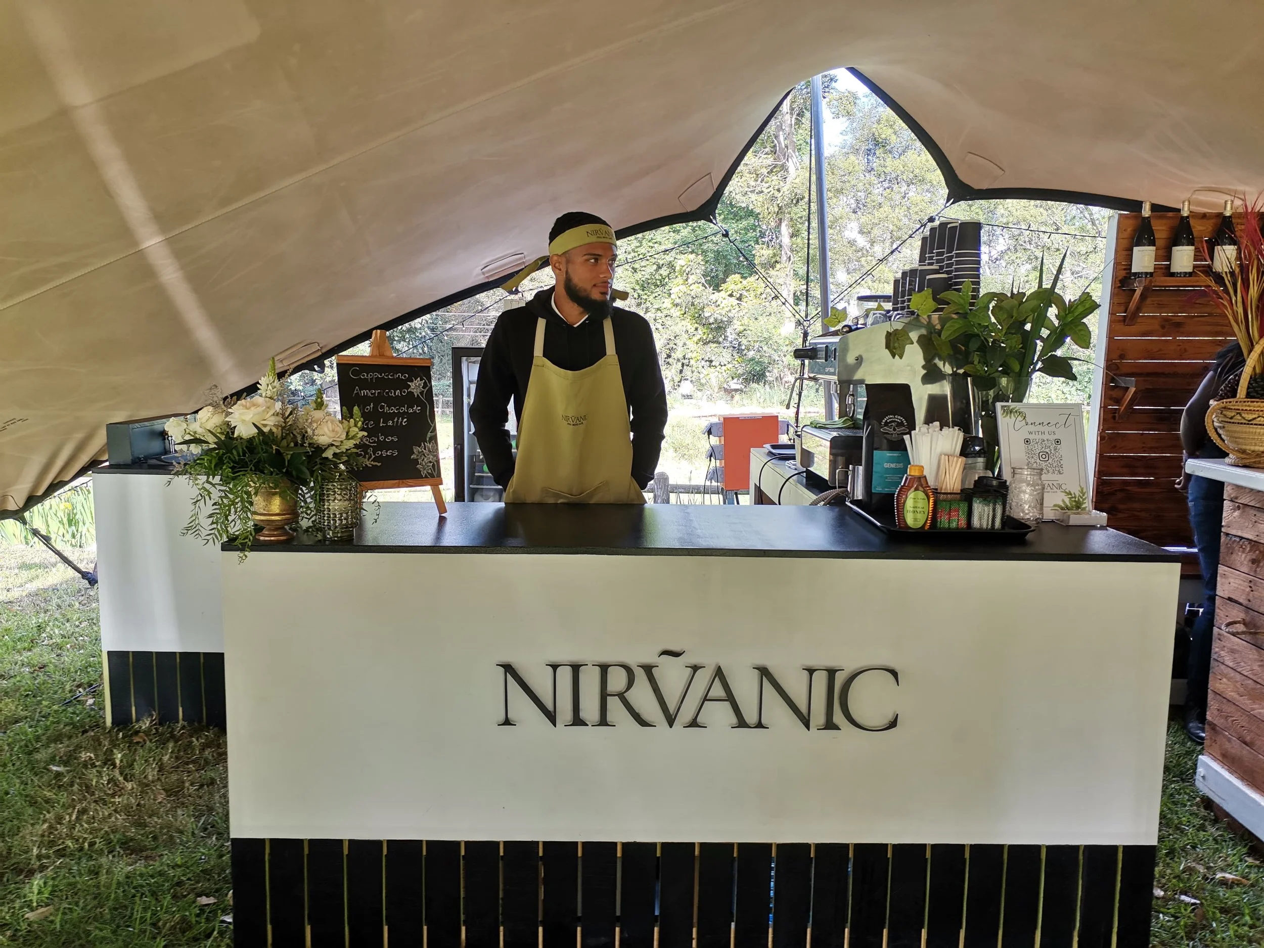 A barista standing behind a coffee stand with a sign labeled 'NIRVĀNIC'. The stand has flowers and a chalkboard menu, with trees visible in the background.