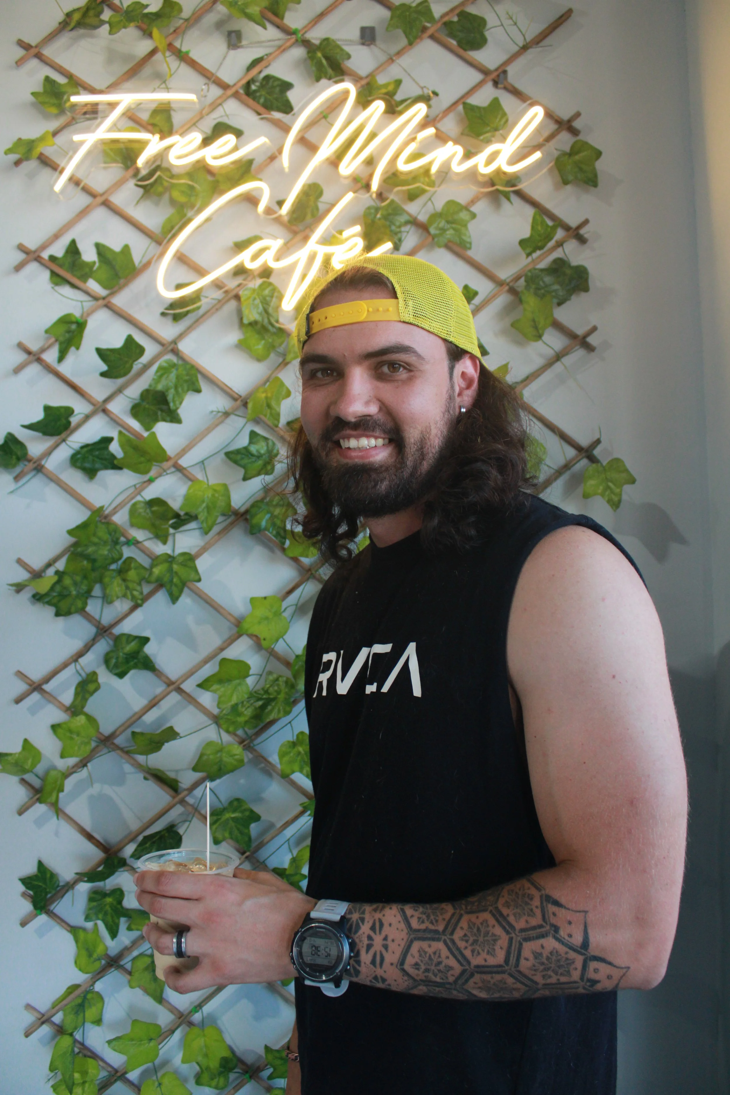 A man with long hair, beard, and tattoos on his arm, wearing a yellow cap backwards and sleeveless black shirt, standing in front of a wall with a green ivy decoration and a neon sign that reads 'Free Mind Café'.