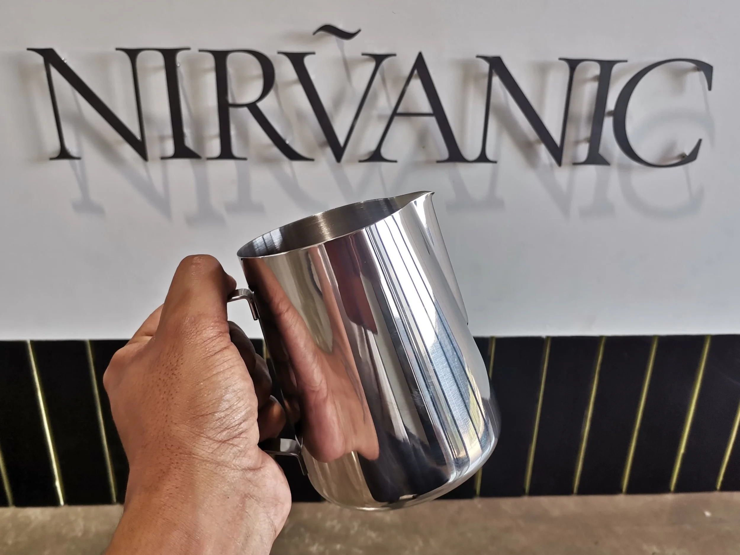 A person's hand holding a shiny metal jug in front of a sign that reads 'NIRVÁNIC' with shadow effects.