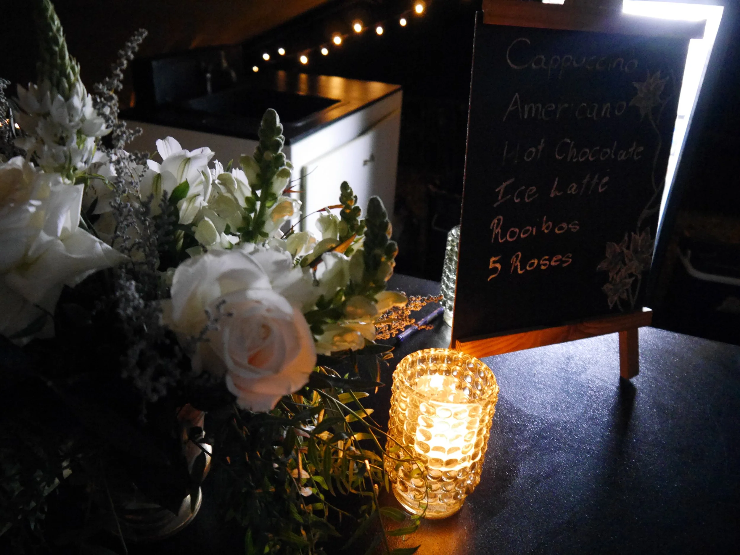 A blackboard menu listing cappuccino, americano, hot chocolate, ice latte, rooibos, and five roses, illuminated by a small illuminated candle, with a bouquet of white flowers nearby.