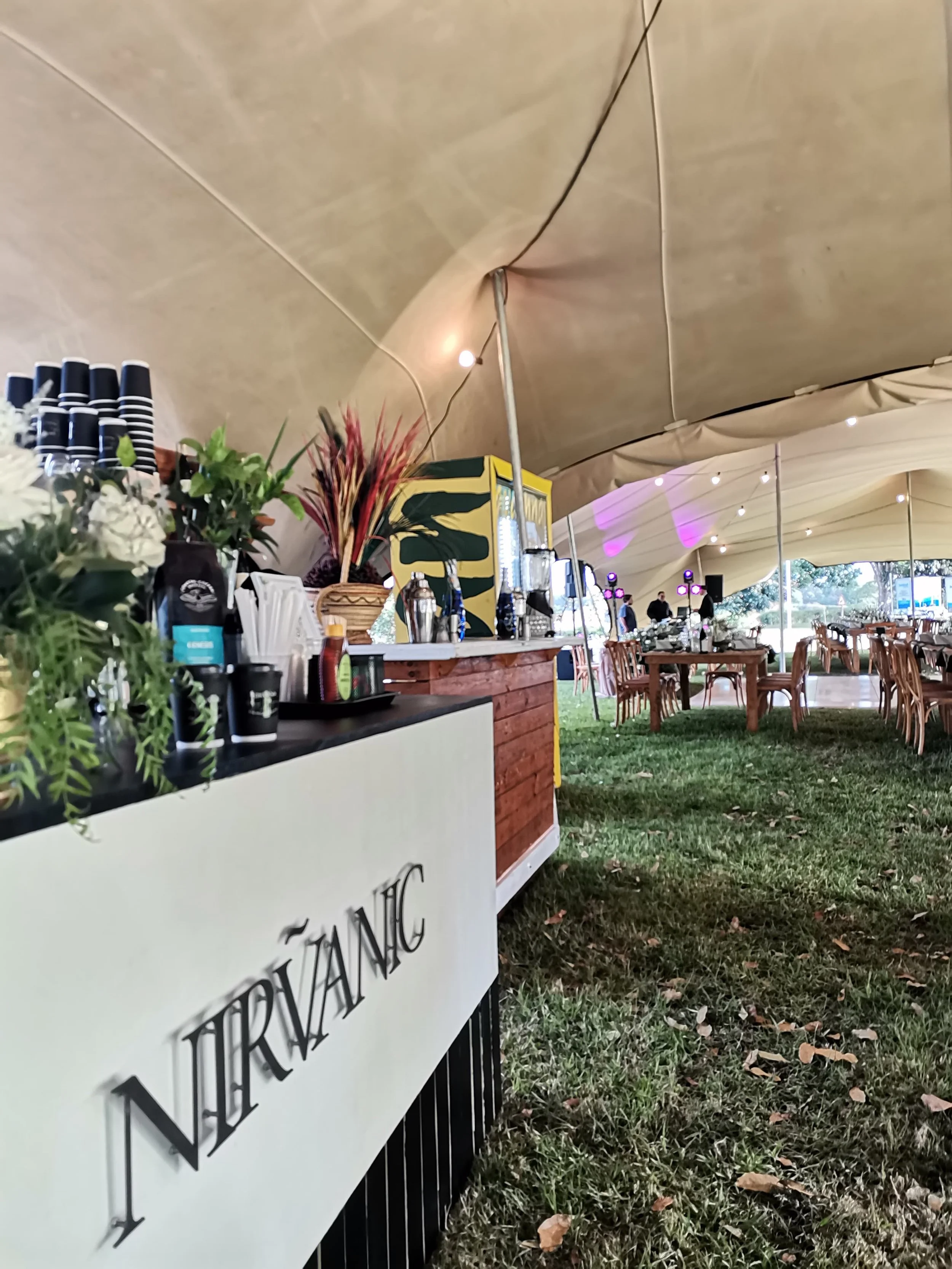 Outdoor event tent with a bar area featuring a white sign that reads 'NIRVÁNIC,' wooden tables and chairs, string lights, and some purple uplighting, with trees visible outside the tent.