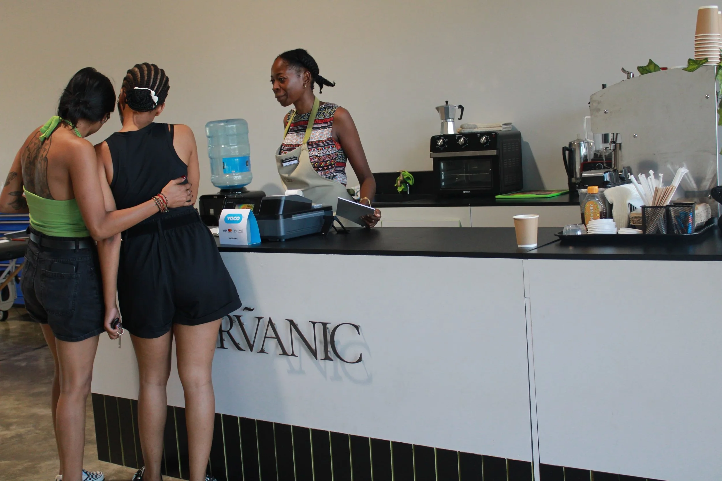 Three women at a cafe counter inside a modern space. Two women are ordering or paying while the third woman is working behind the counter. The counter has a water dispenser, a cash register, coffee machine, and disposable cups.