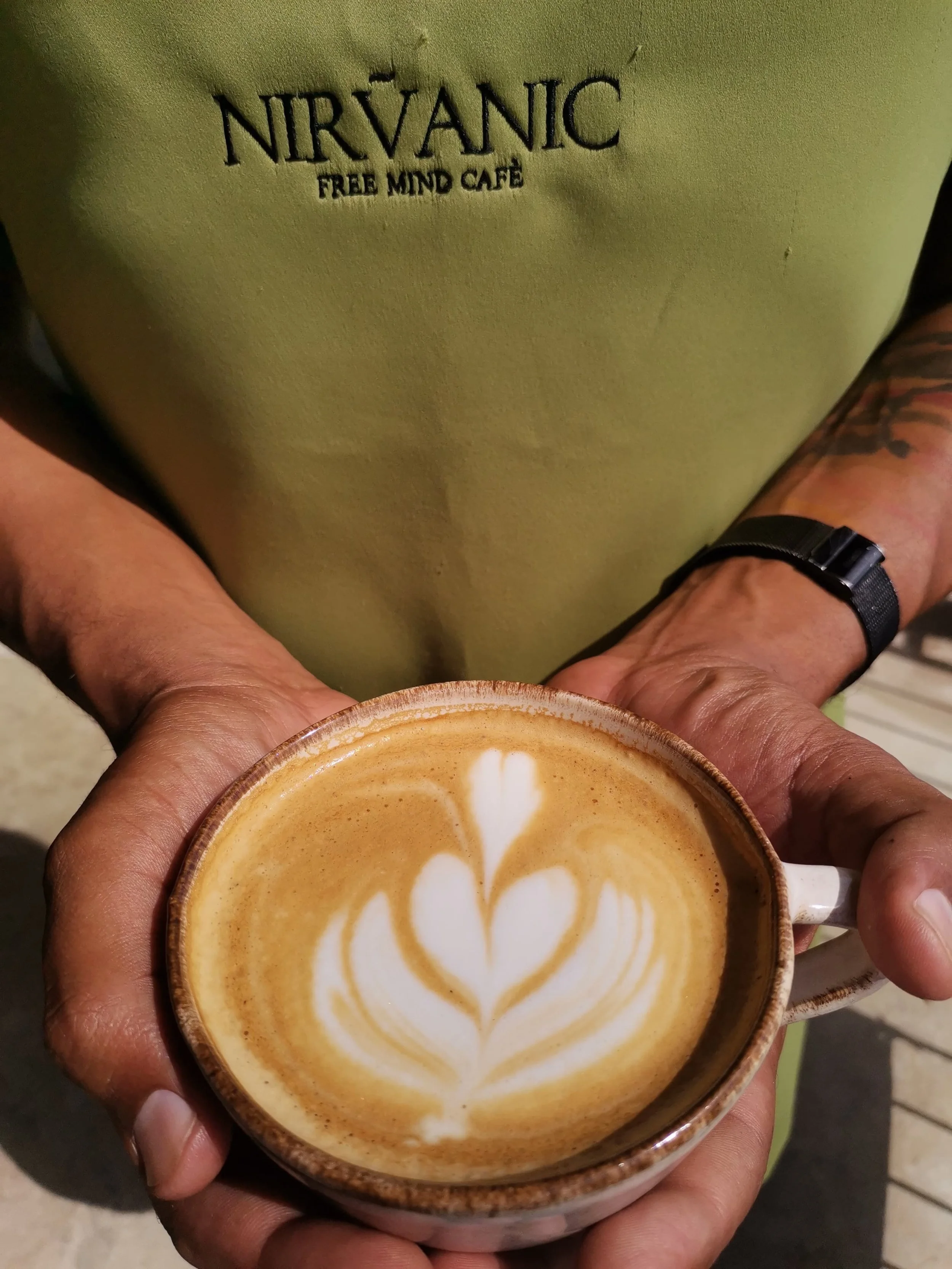 Person holding a ceramic cup of latte art coffee with a leaf design, wearing a green Nirvāṇic free mind cafe t-shirt and a black watch.