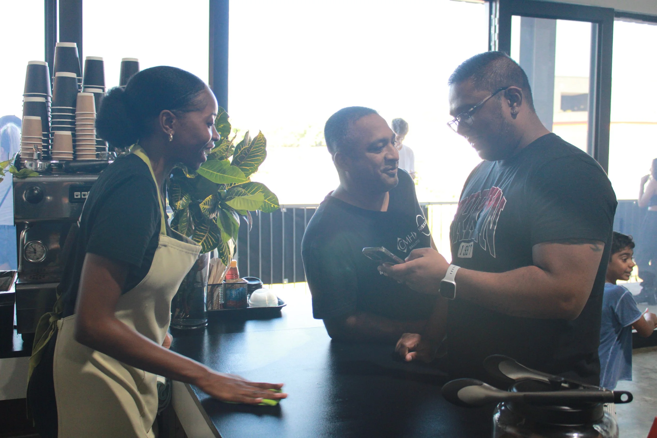 A barista in a beige apron cleaning a counter while talking to a customer at a coffee shop. The customer and another man are looking at a phone together and smiling. There are cups and a coffee machine in the background.