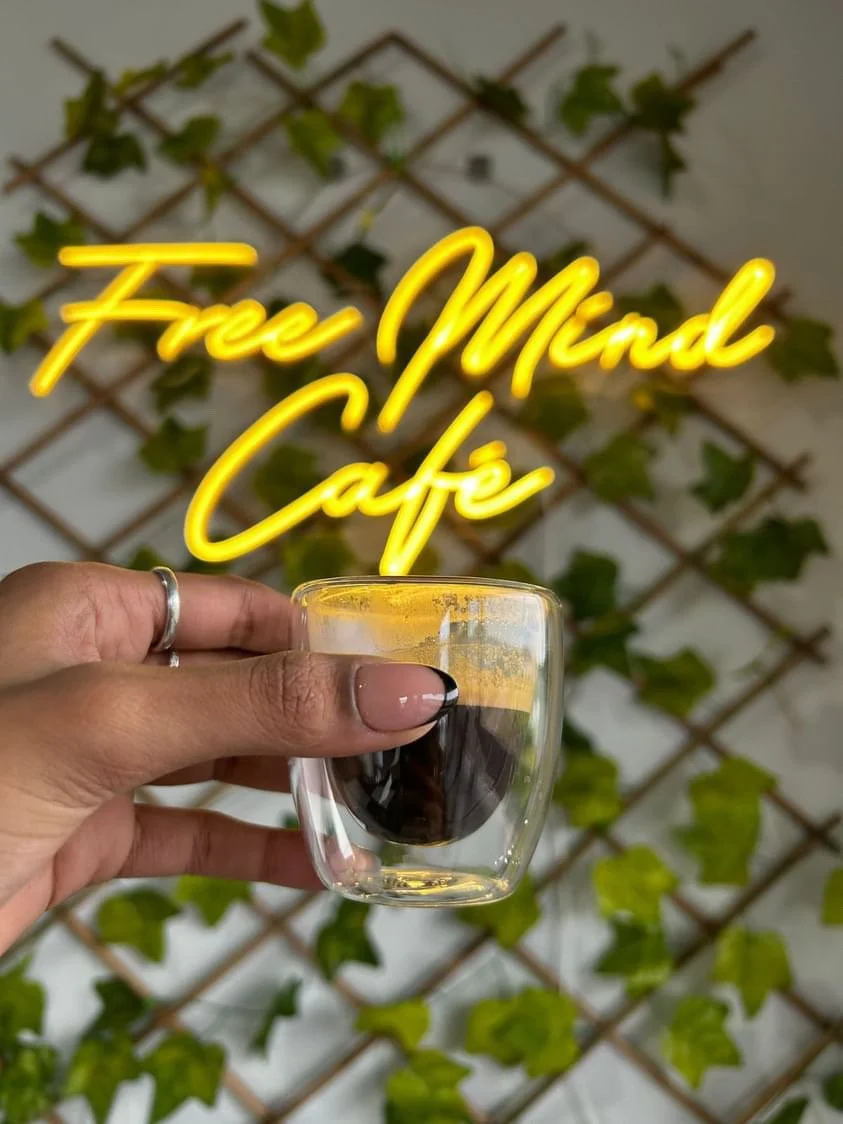 A hand holding a clear glass cup with espresso black coffee. In the background, a yellow neon sign says 'Free Mind Café' and a decorative wooden lattice with green ivy leaves.