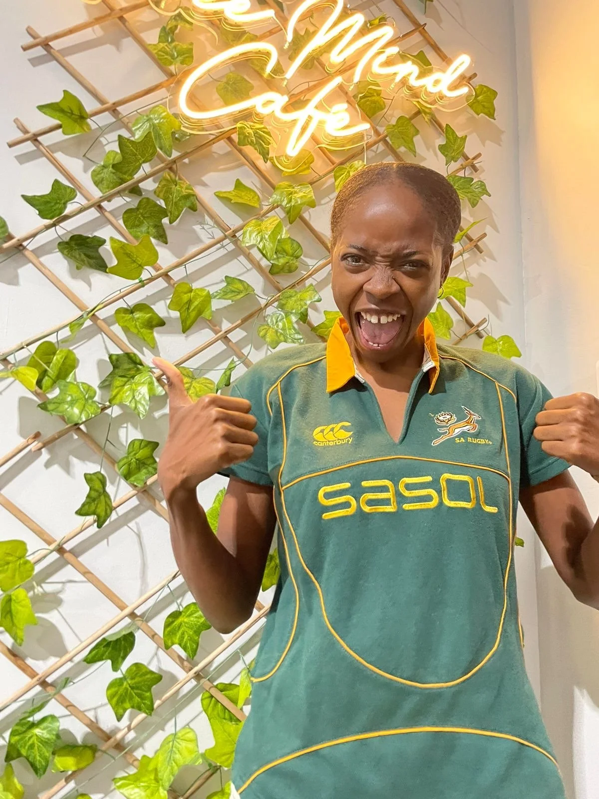 A woman wearing a green Rugby shirt with yellow accents, standing in front of a wall with green ivy and a neon sign that reads 'Free Mind Cafe'. She is making a happy, excited expression, with fists clenched in a celebratory pose.