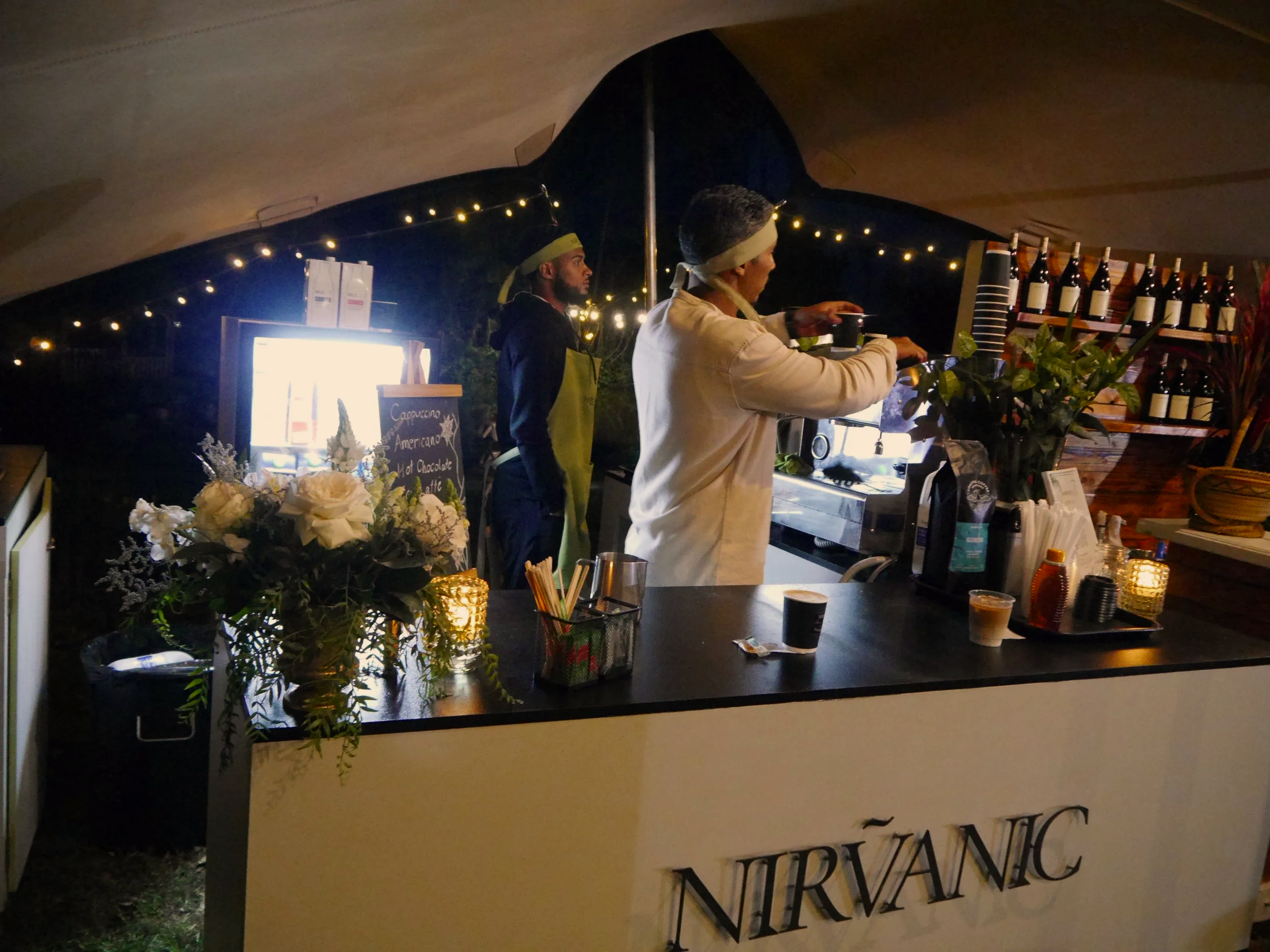 Two people working behind a coffee bar at Nirvanic, with bottles and flowers on the counter, and string lights overhead.