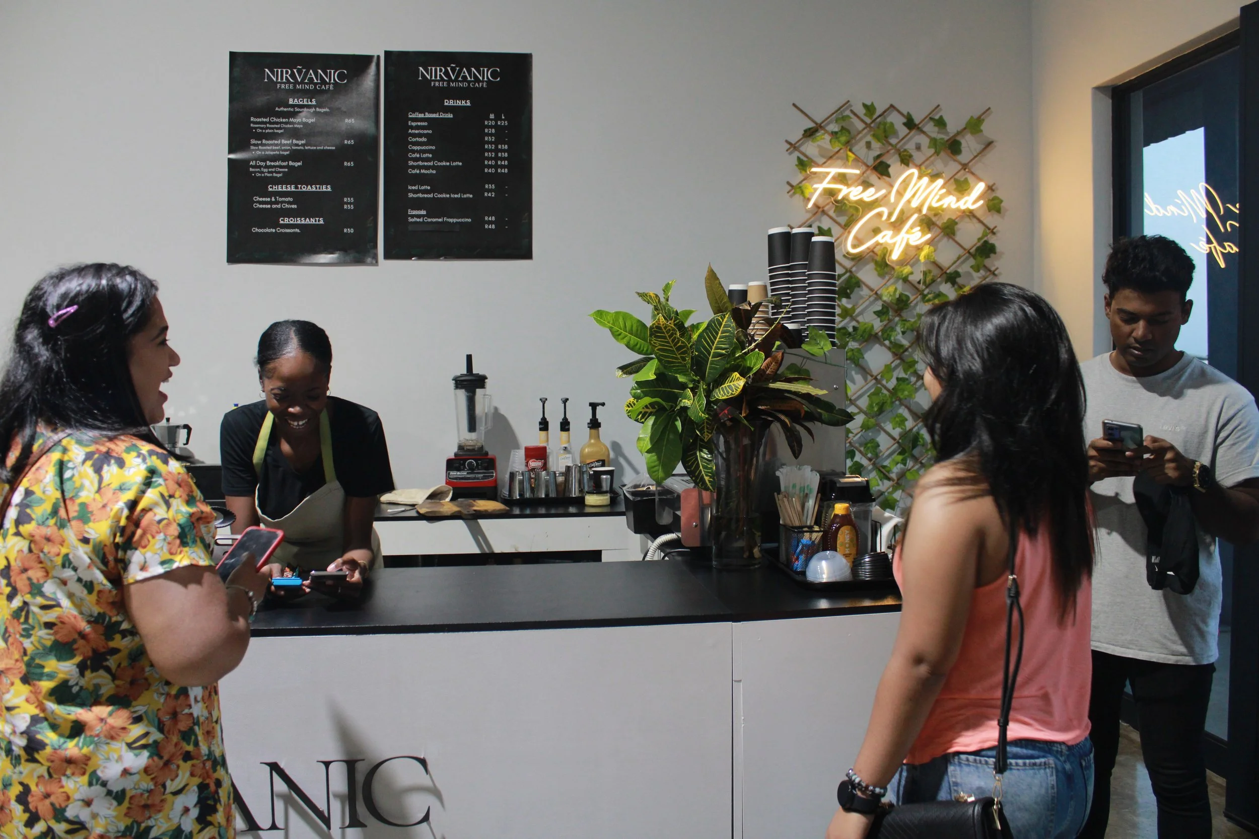 People standing at a counter in a cafe with a neon sign that says 'Free Mind Cafe', a menu on the wall, and a staff member behind the counter preparing orders.