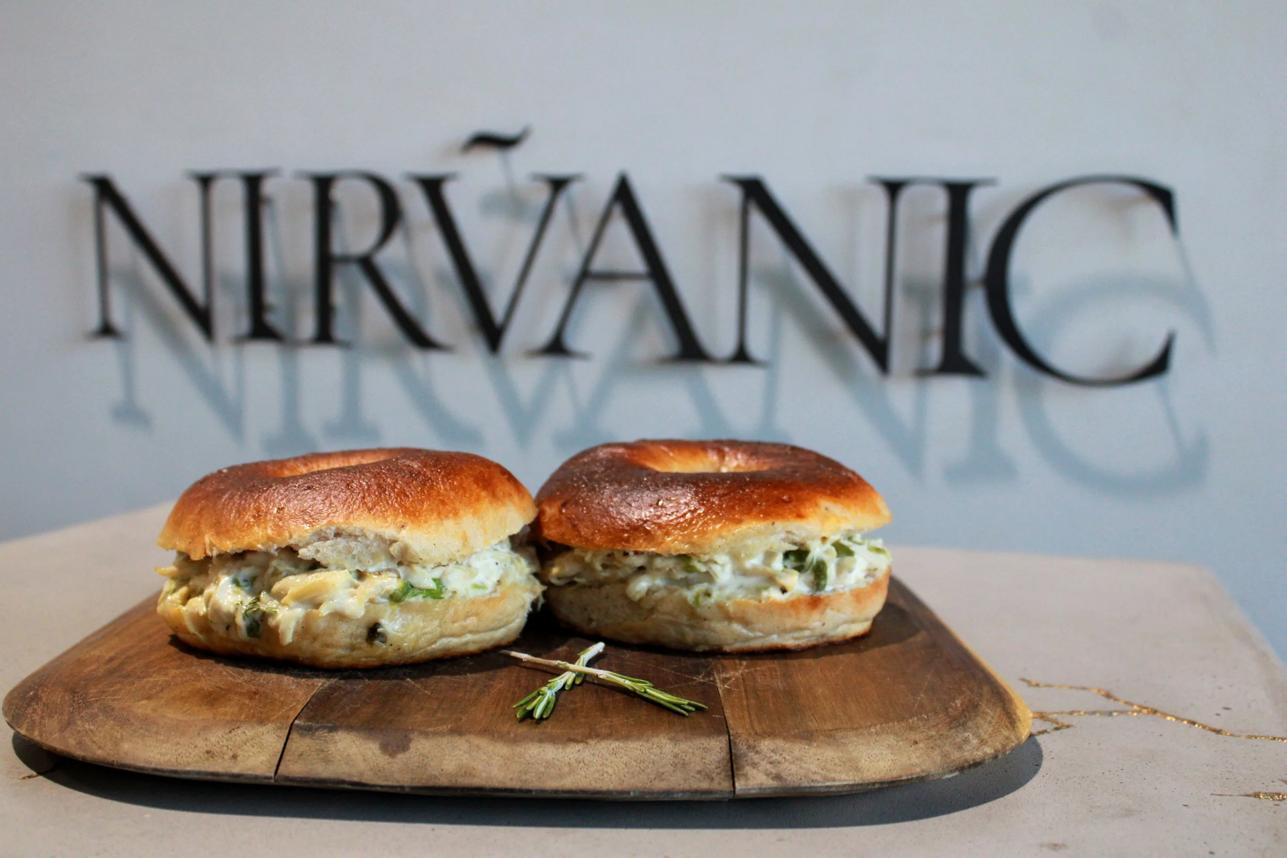 Two bagels with chicken & mayo and rosemary herbs on a wooden board in front of a wall with the word "NIRVANIC" in large black letters, casting shadows.