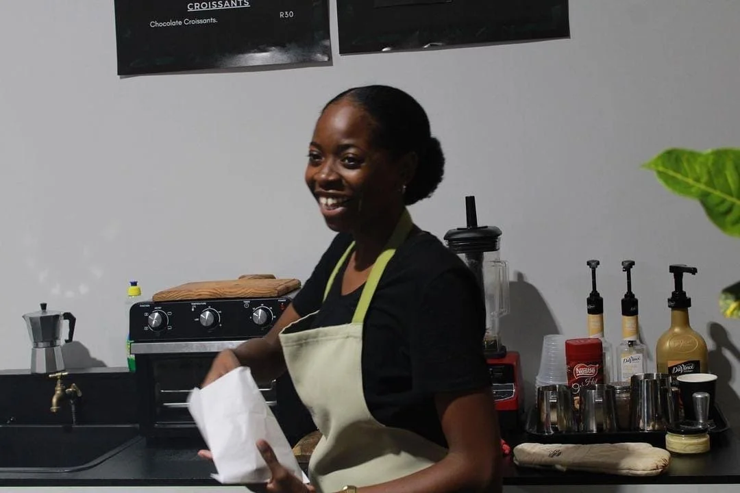 A smiling woman in a black shirt and beige apron standing behind a coffee counter, with various coffee equipment and supplies in the background.