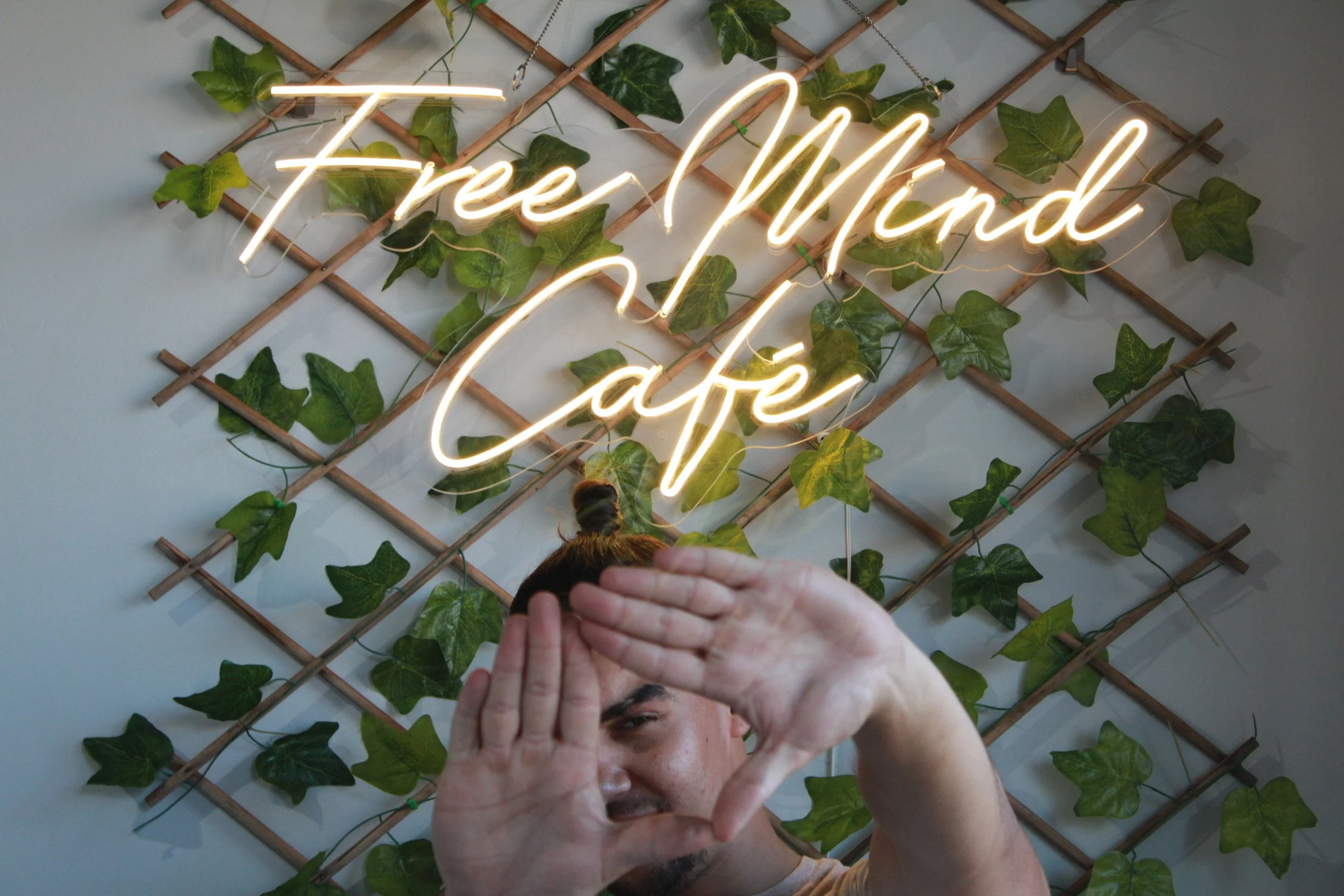 Person with a top knot hairstyle making a heart shape with hands in front of a wall with a neon sign that reads 'Free Mind Café' and decorative green ivy leaves.
