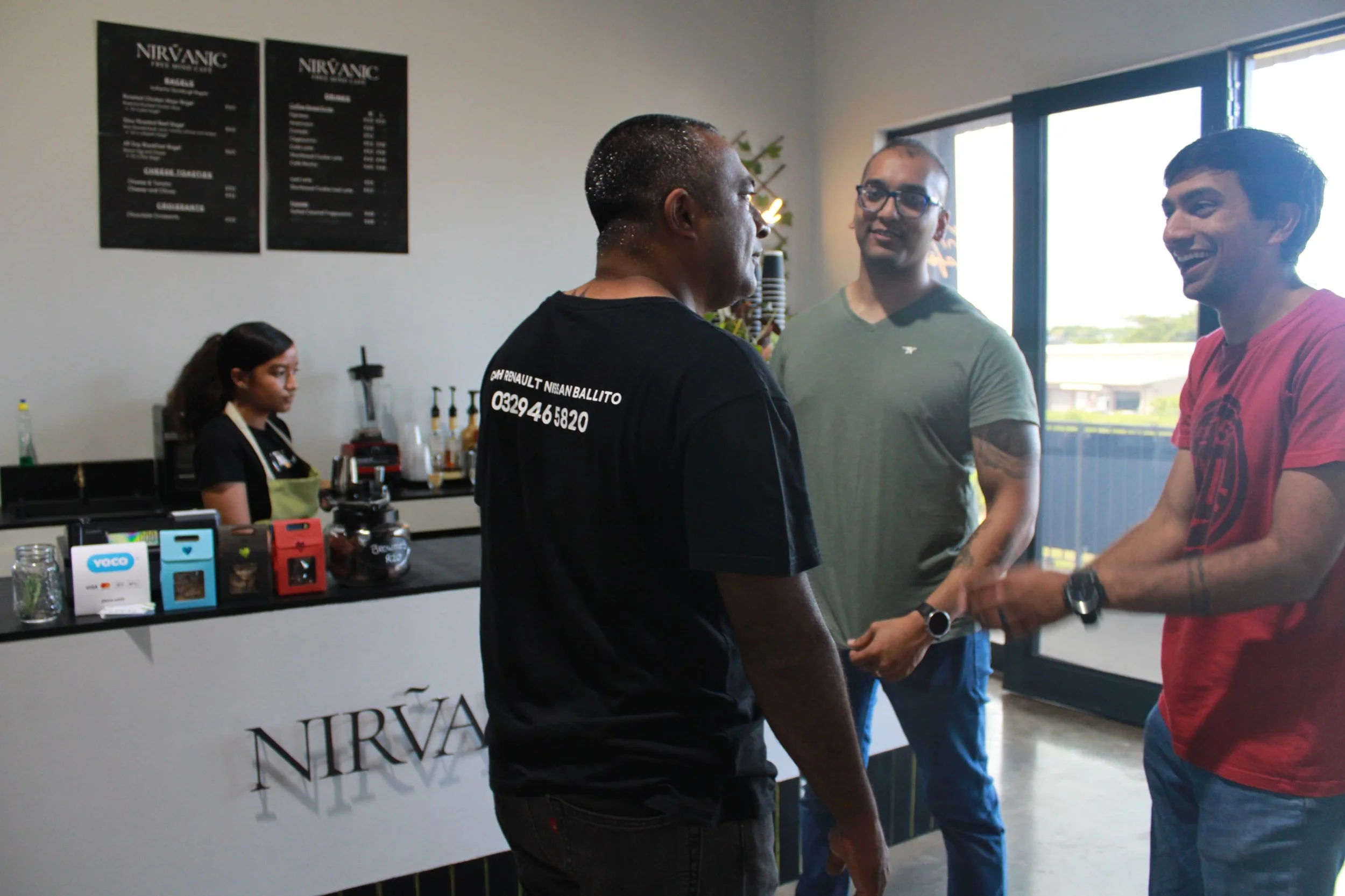 Three men are smiling and talking to each other inside a coffee shop. Two are facing forward, and one is turned sideways, all engaging in conversation. Behind them, a woman is preparing drinks behind the counter, which displays a Nirv sign and menu b