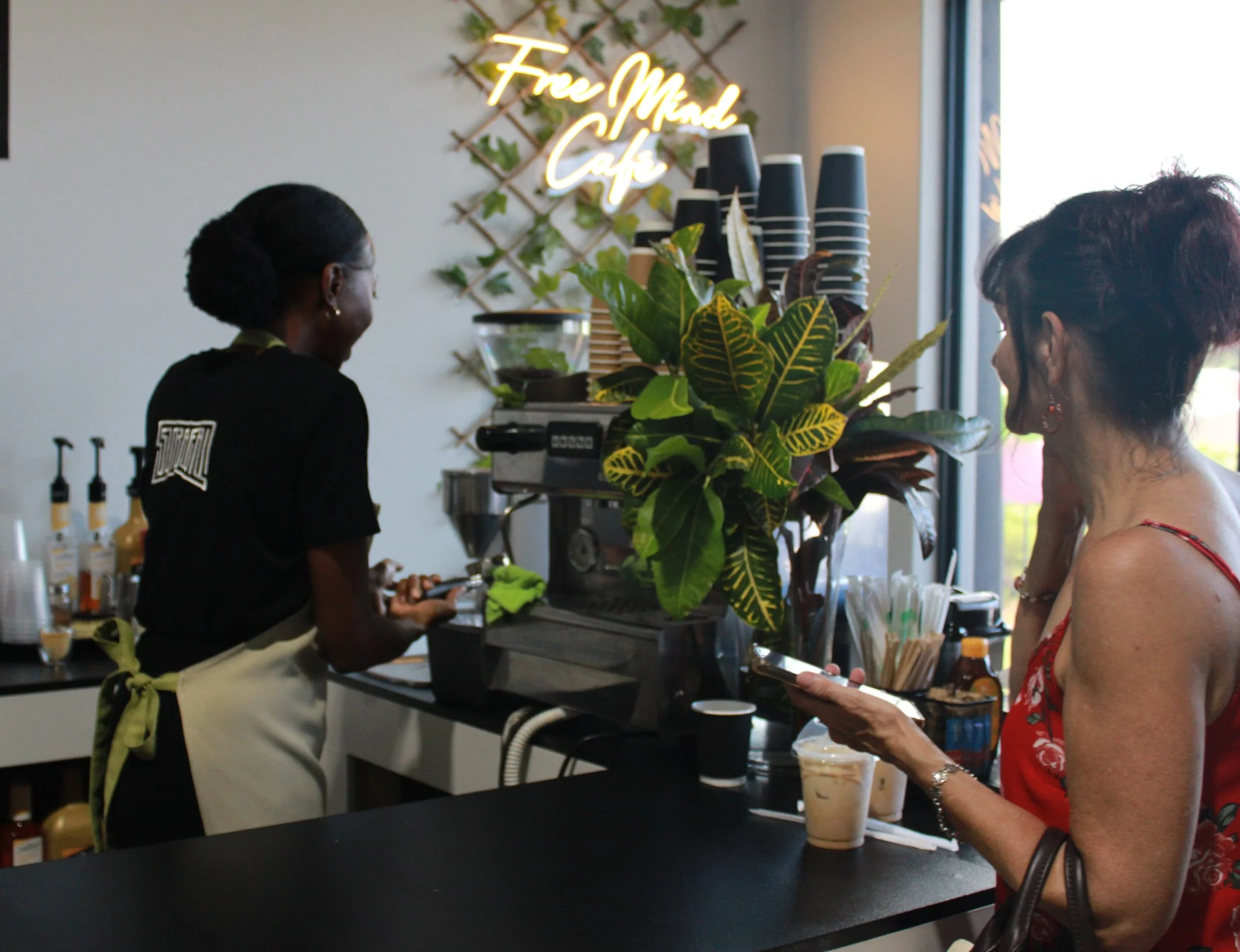 A barista and a customer at a coffee shop with a neon sign that reads 'Free Mind Cafe'. The customer is holding a phone and wearing a red dress, while the barista is wearing a black shirt and light-colored apron. There is a large green plant on the c