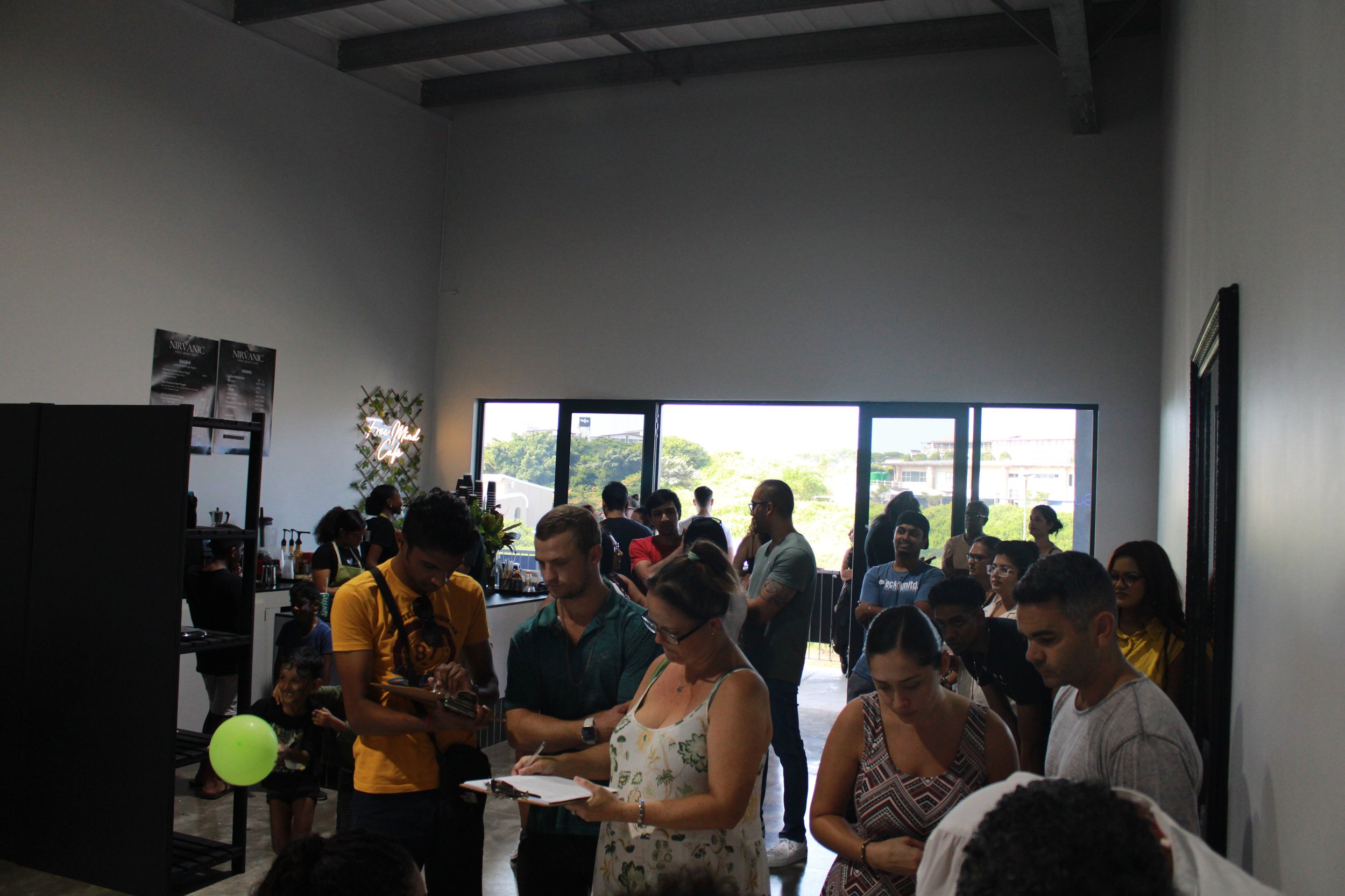Indoor gathering of people at a social event or cafe, with some standing in line, some talking, and others using their phones, with large windows in the background showing trees and buildings.