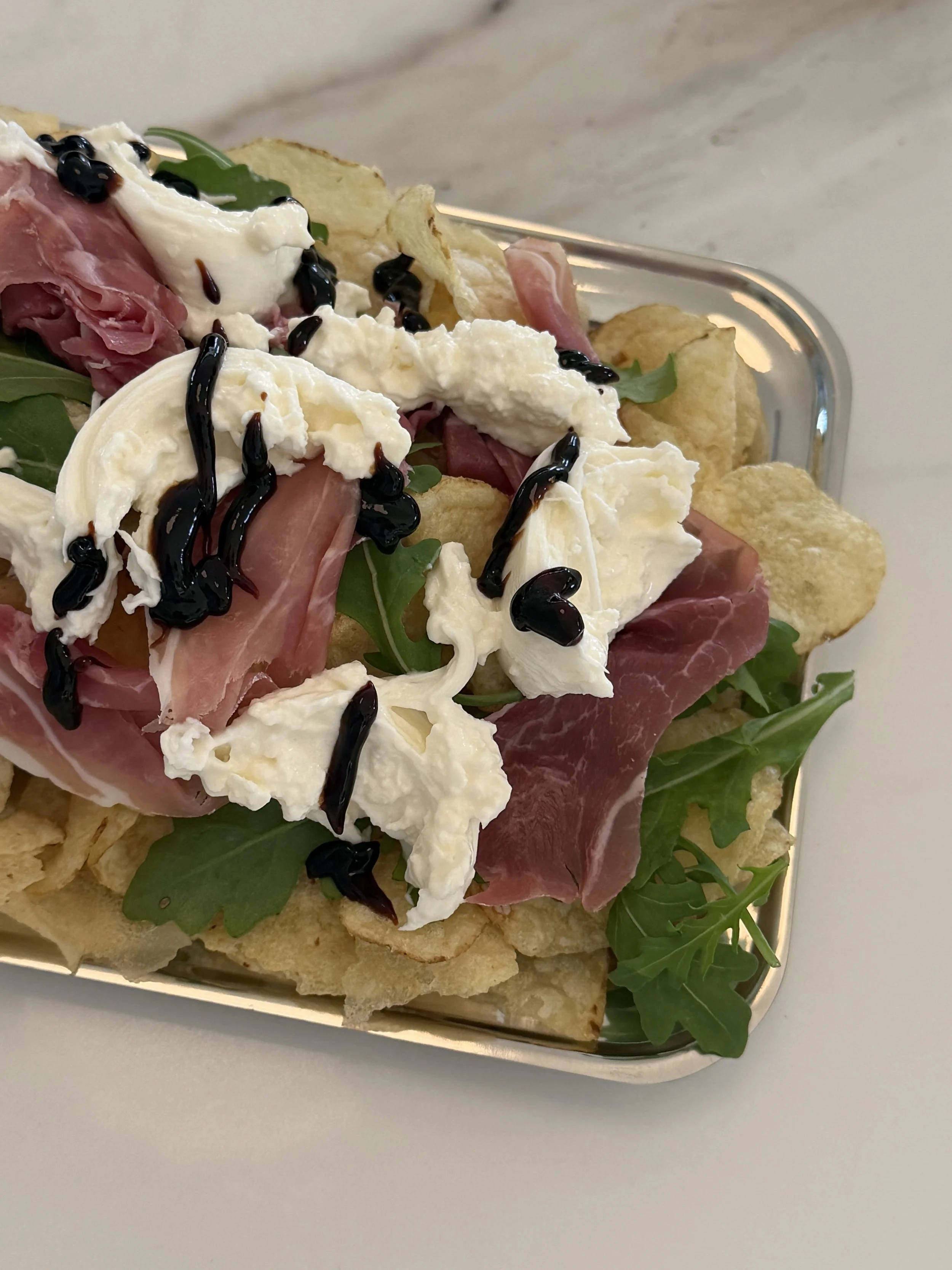 A glass dish with chips, arugula, prosciutto, mozzarella cheese, and balsamic glaze.