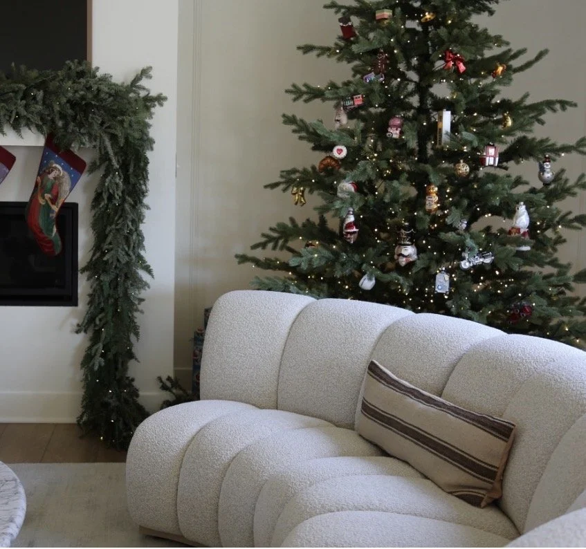 A decorated Christmas tree with ornaments and lights, a festive fireplace mantel with stockings, and a cozy beige sofa with a striped pillow in a living room.