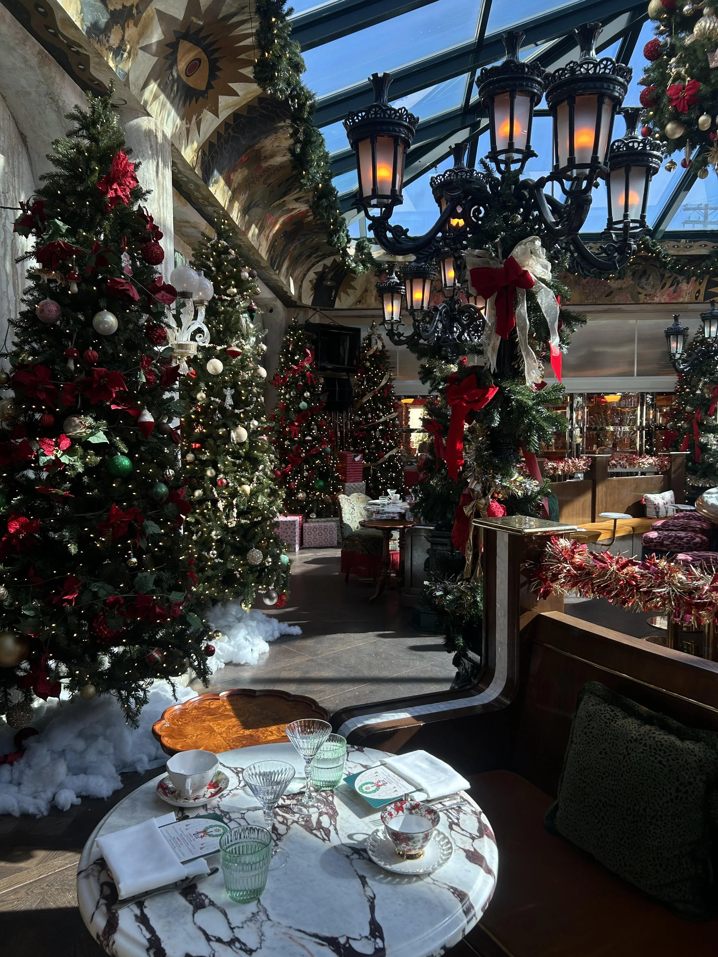 Festively decorated indoor space with Christmas trees, garlands, and wrapped gifts, featuring a table set with dishes, glasses, and napkins, under a glass ceiling with a large ornate chandelier.