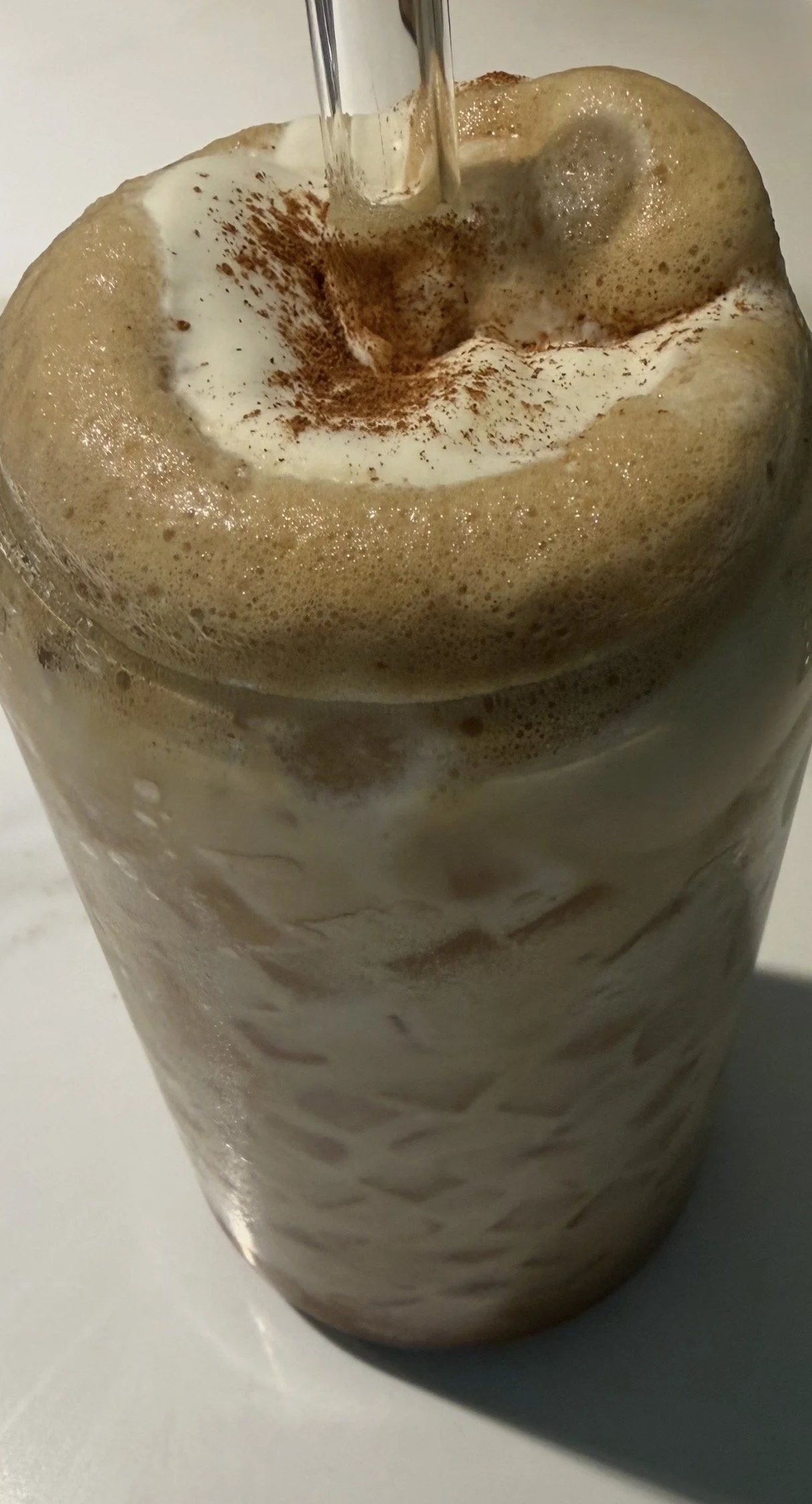 A tall glass of iced coffee topped with whipped cream and a dusting of cocoa powder. A straw is inserted into the drink.