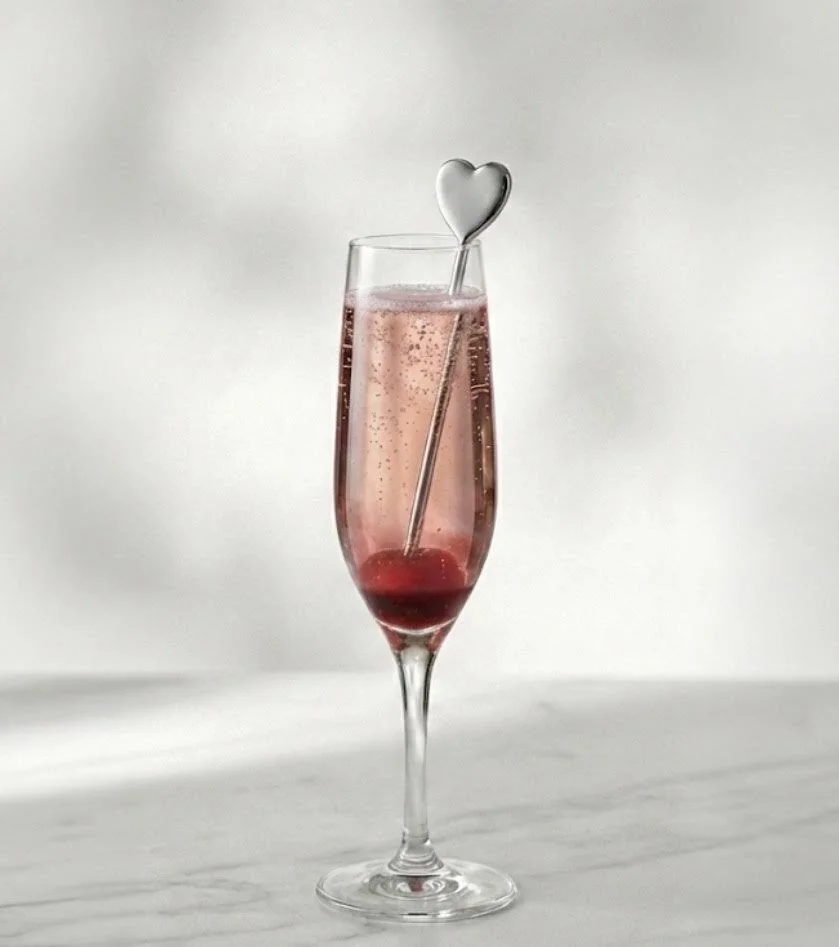 A tall champagne flute filled with pink bubbly liquid, garnished with a red cherry at the bottom and a heart-shaped stir stick.