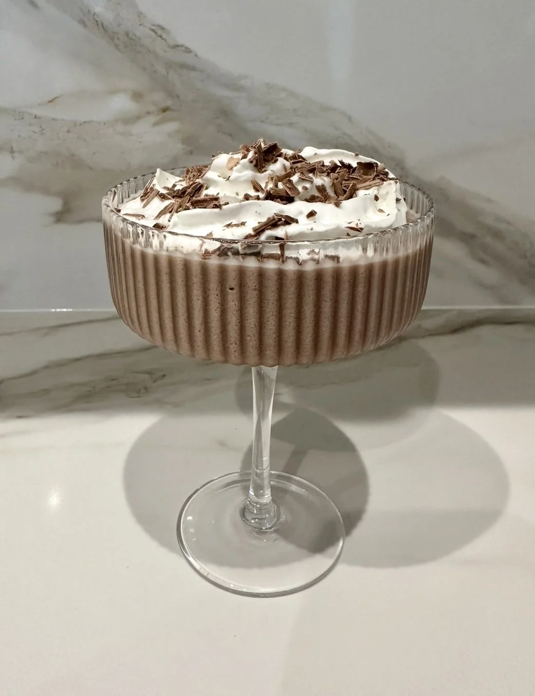 Chocolate mousse dessert topped with whipped cream and chocolate shavings in a decorative glass on a white marble background.