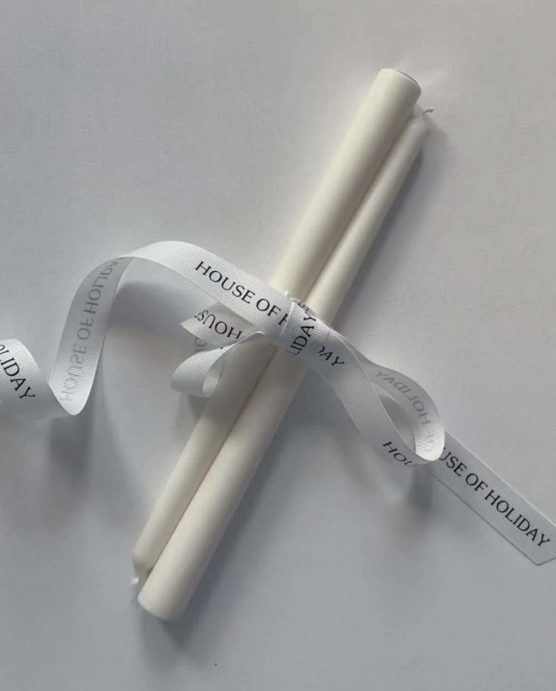 Two white candles tied with a white ribbon labeled 'House of Holiday' on a plain white surface.