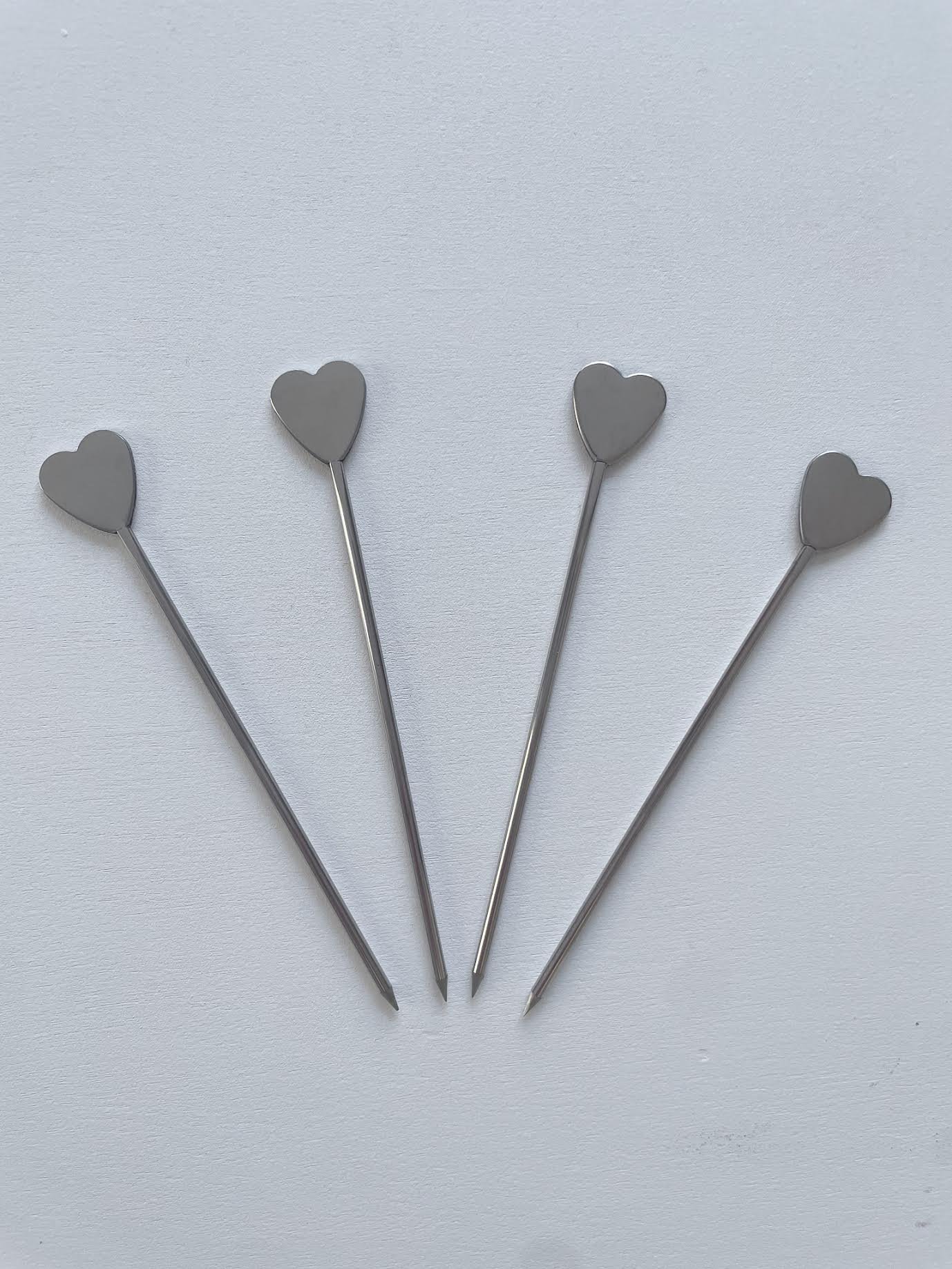 Four silver metal cocktail picks with heart-shaped tops arranged on a white surface.