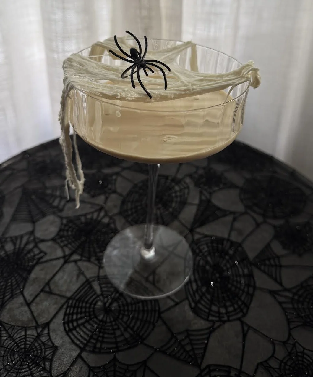A Halloween-themed cocktail in a coupe glass decorated with fake spider web and a plastic black spider on top, placed on a table with spiderweb-patterned tablecloth.