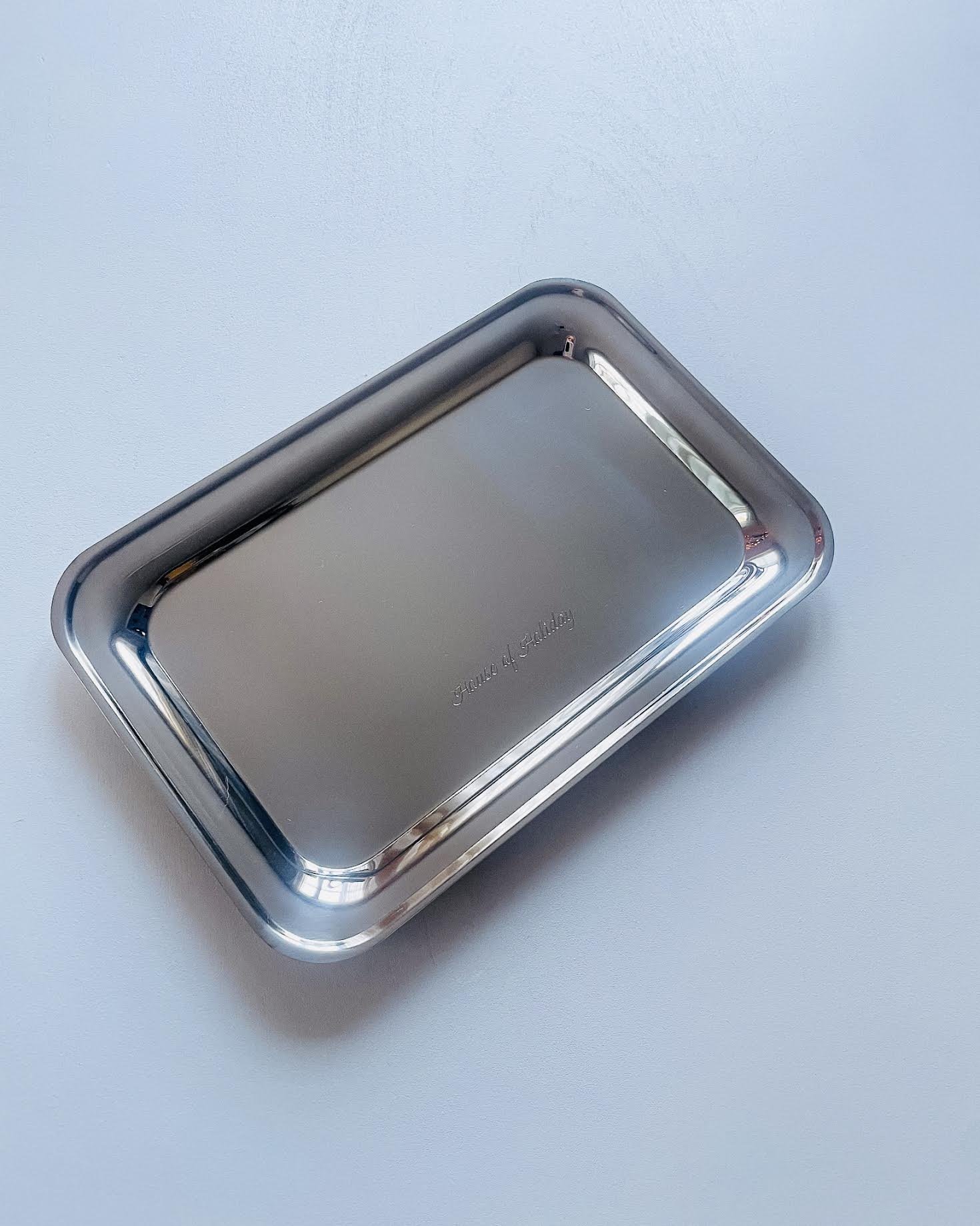 Metallic rectangular tray with rounded edges on a light blue surface, with faint engraved text in the center.