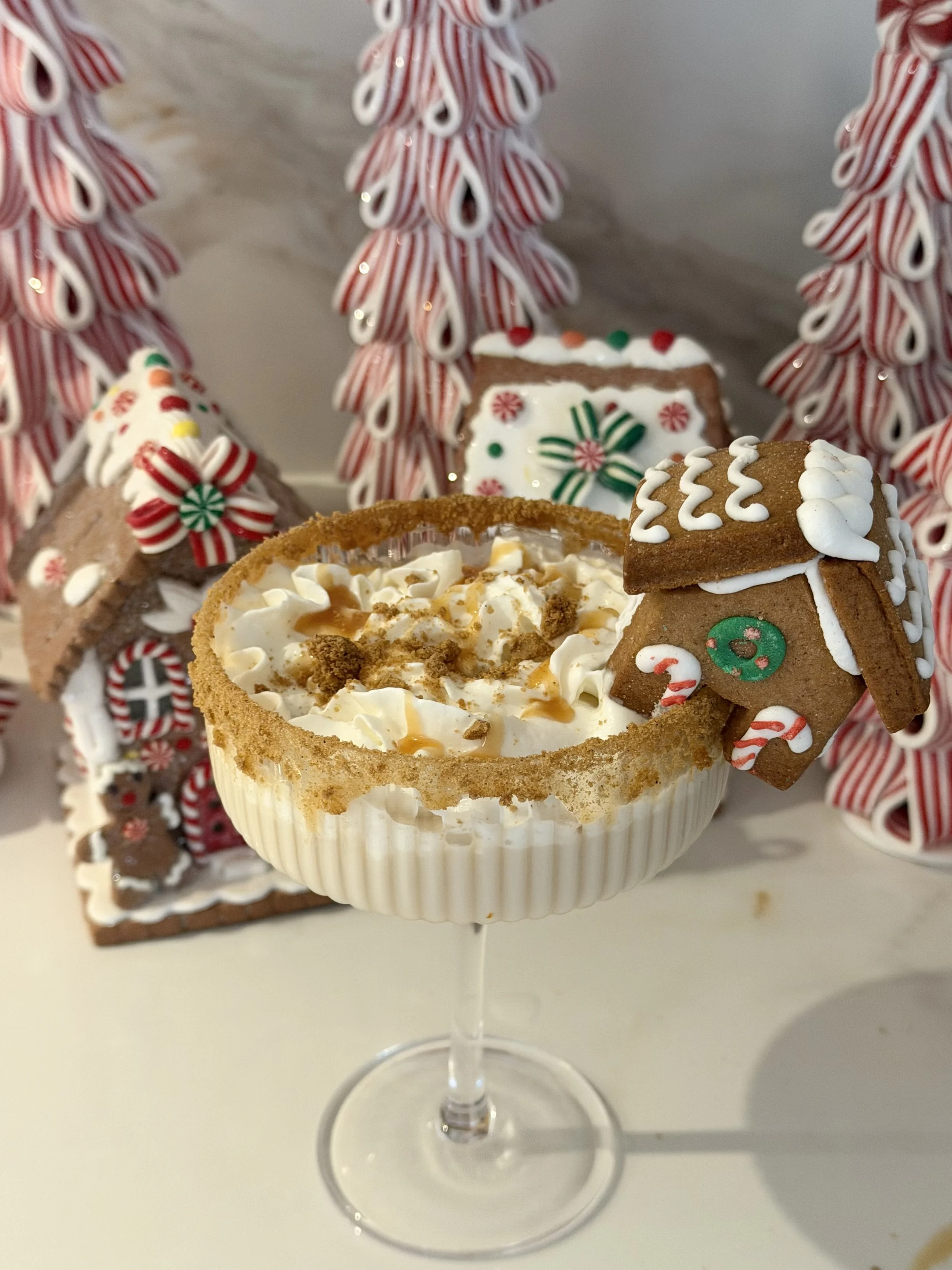 A festive holiday dessert in a stemmed white bowl with whipped cream, caramel drizzle, and crumbled cookies, decorated with a gingerbread house cookie and peppermint candies, with candy cane and Weihnachtswapen gingerbread cookies and peppermint decorations in the background.